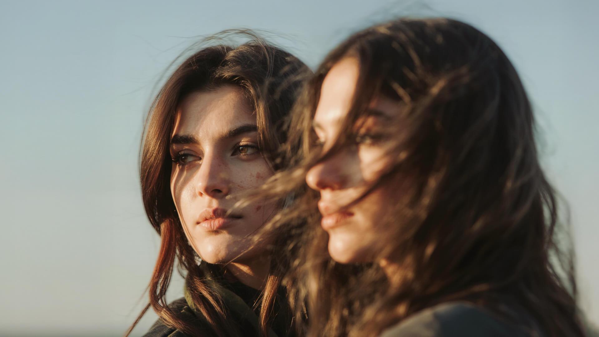Two women with long, dark hair look into the distance, with the golden sunlight highlighting their features against a clear sky.