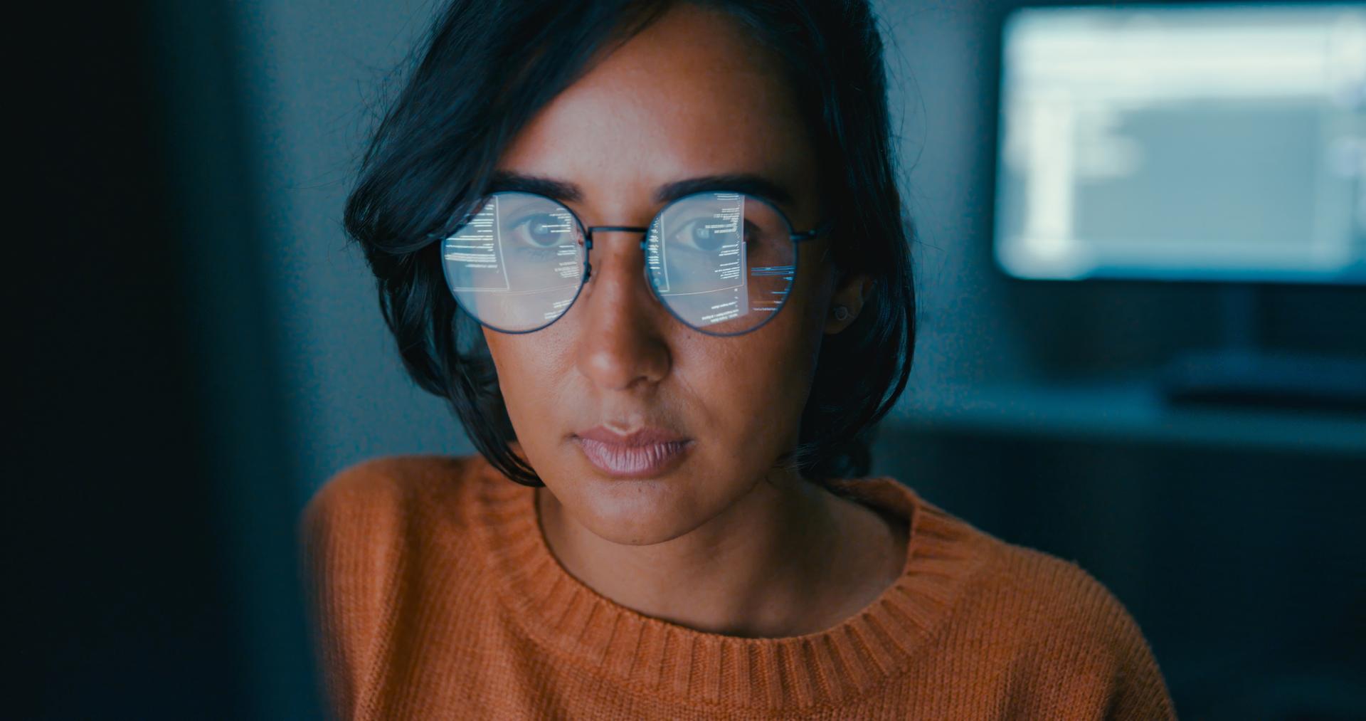 Person wearing glasses, focused on a computer screen with code reflecting in the lenses, in a dimly lit room.