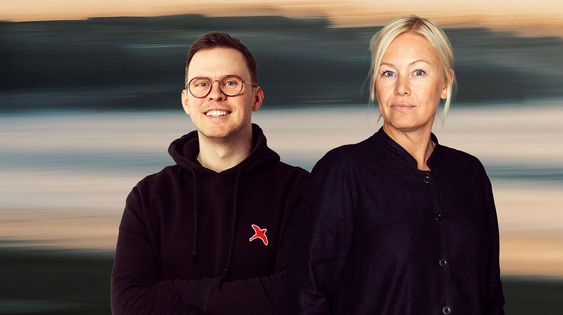 A man in glasses and a woman with blonde hair stand side by side, smiling against a blurred outdoor background.