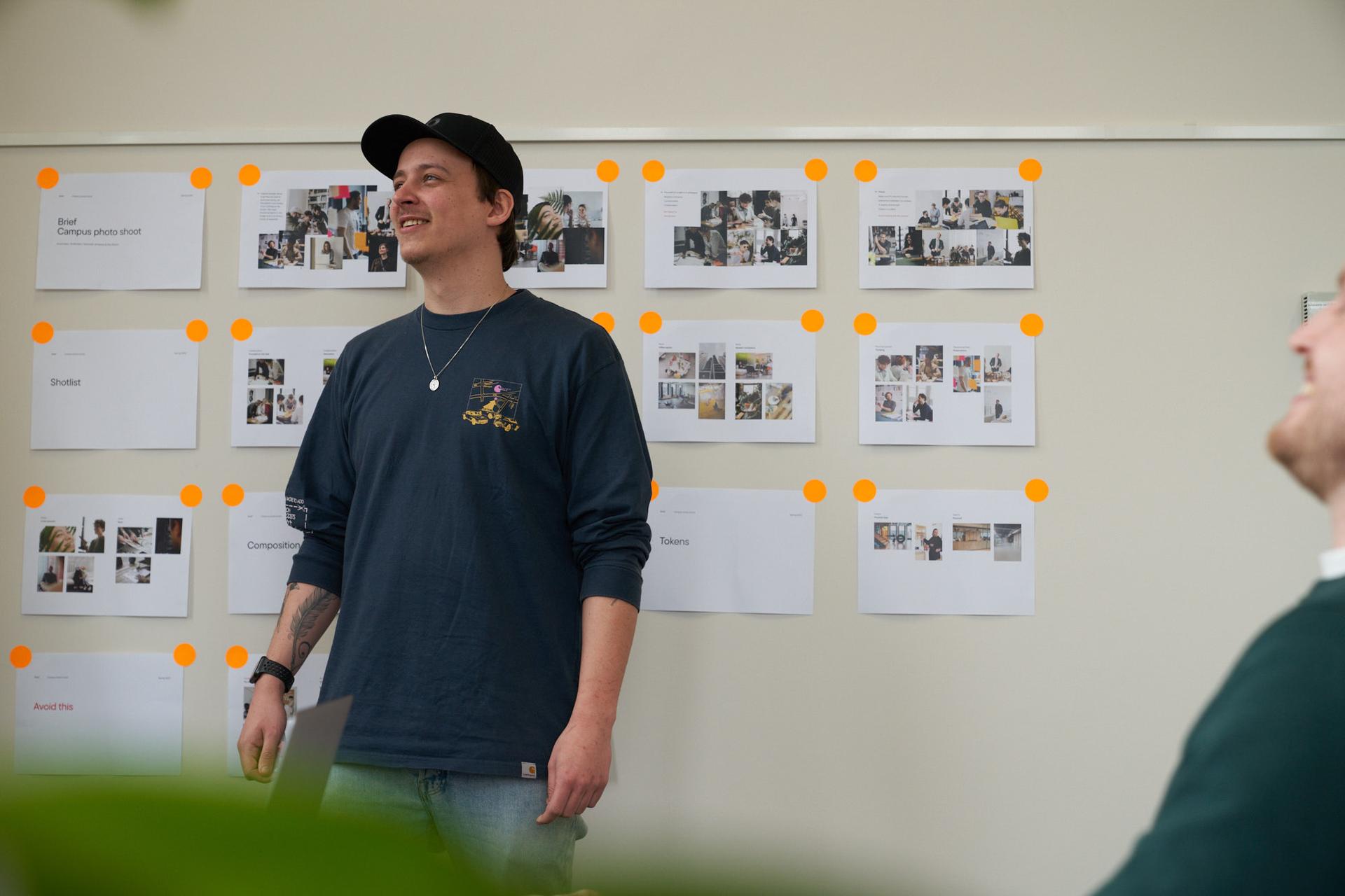 Person in a blue shirt and cap stands smiling in front of a wall with photos and orange dots, while another person is partially visible.