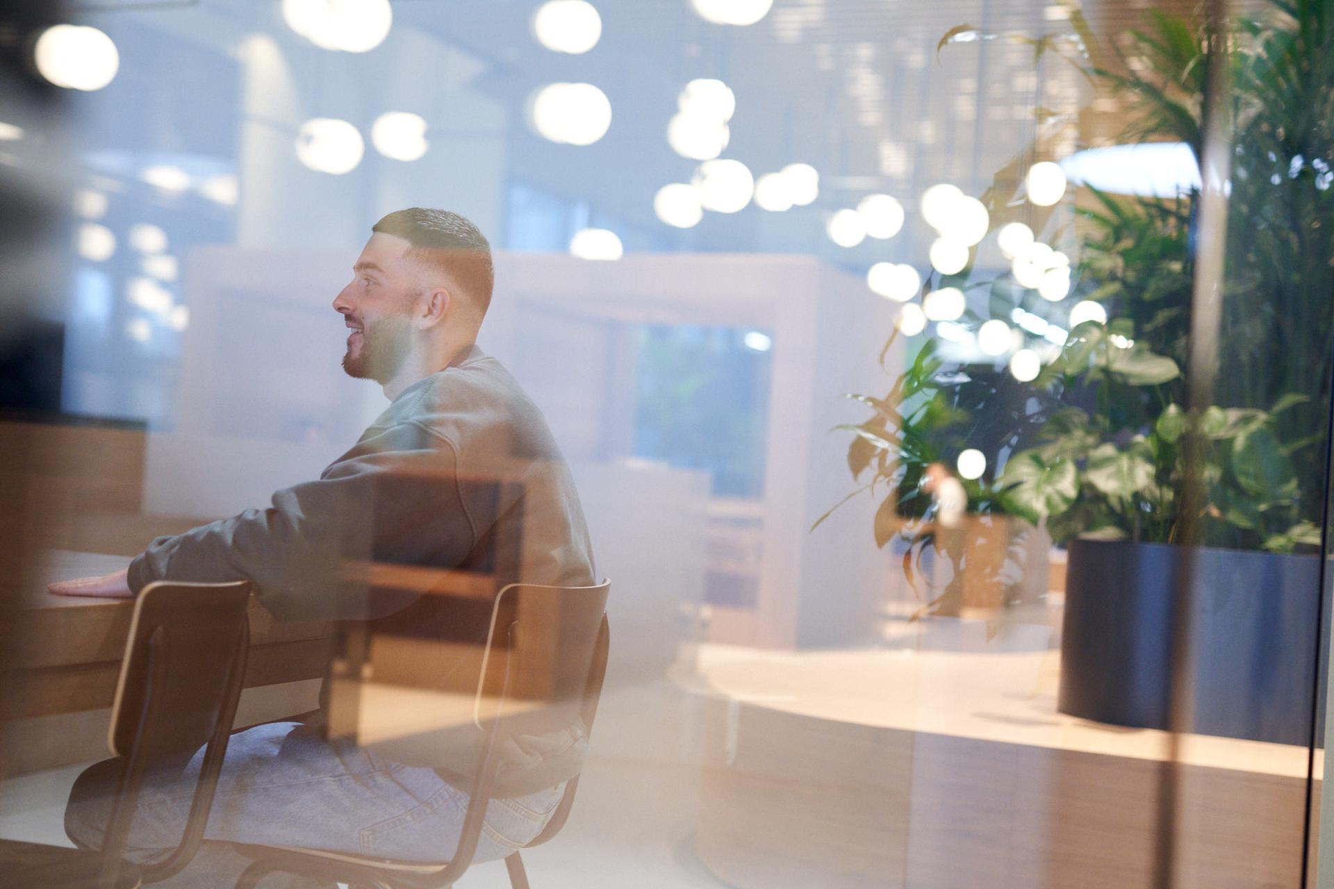 Man sitting at a table in a modern, well-lit office space, with reflections and plants visible through glass.