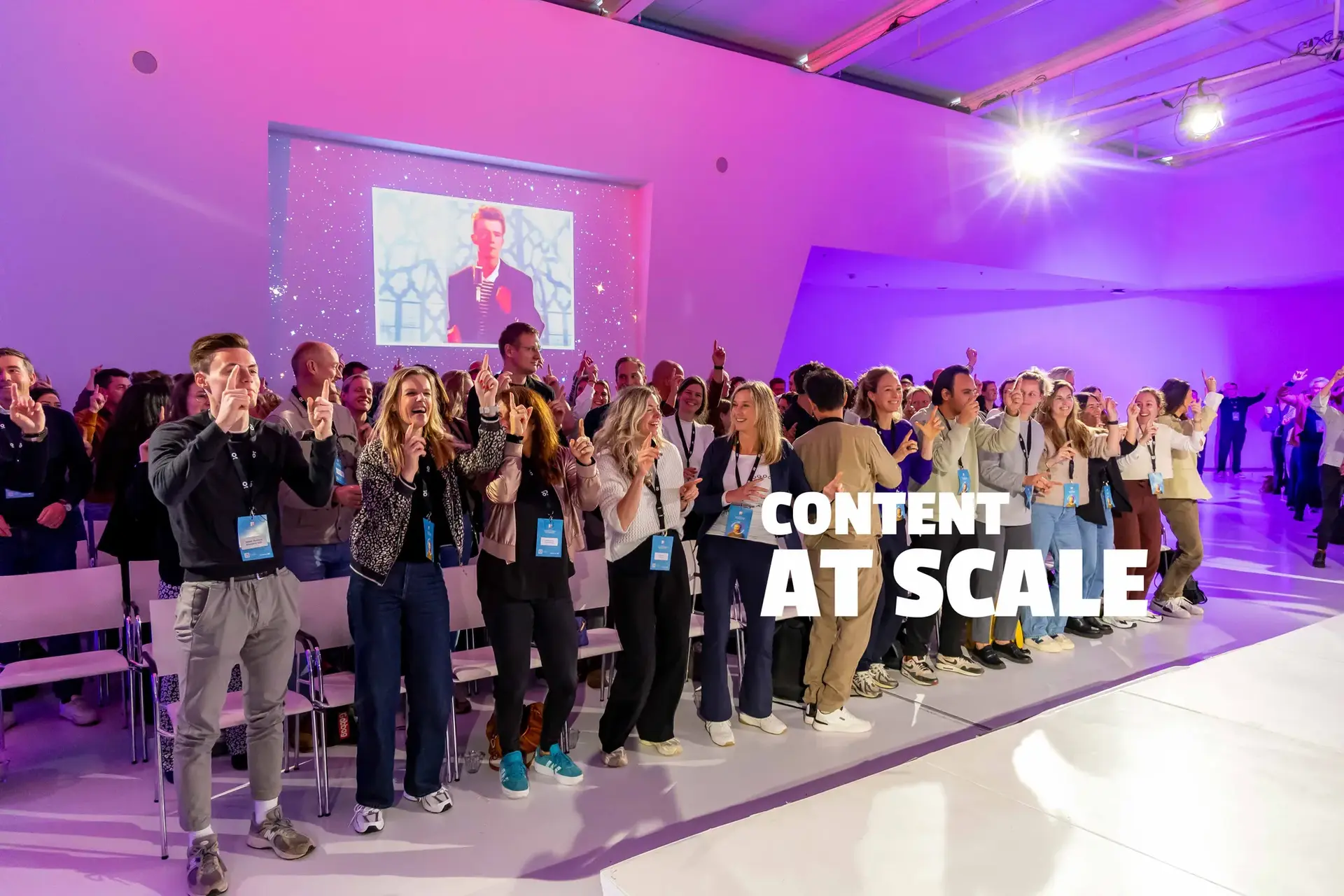 Audience clapping in a modern, purple-lit auditorium with "CONTENT AT SCALE" text projected on the wall.