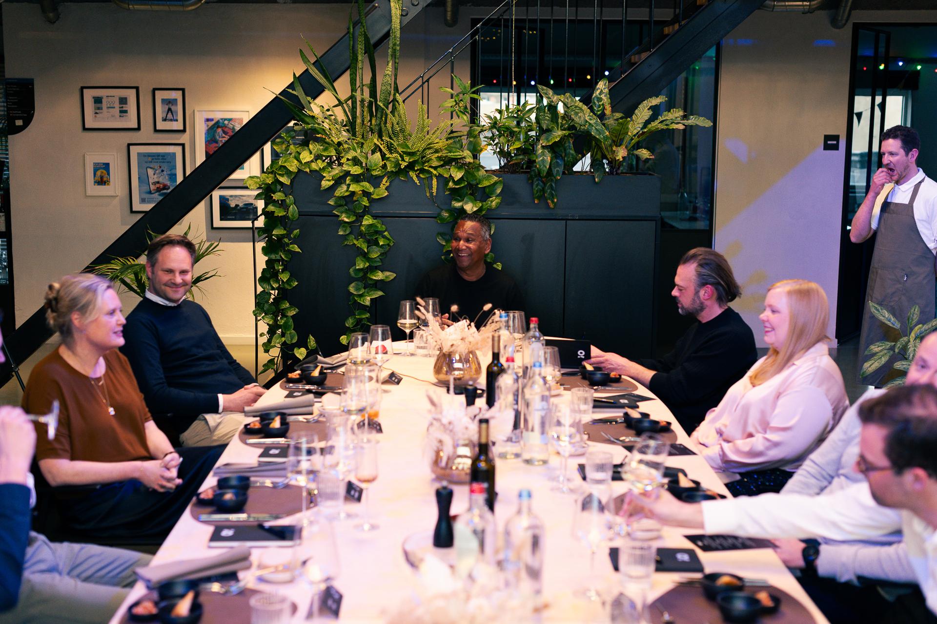 A group of people sitting around a dining table with drinks, plants in the background, and a chef standing near the staircase.