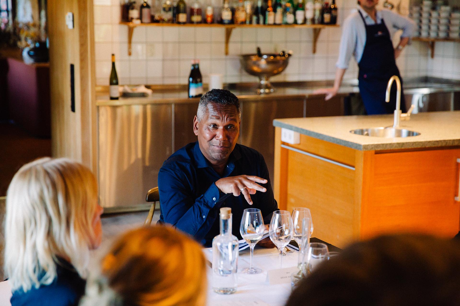 A man in a blue shirt is speaking at a table with wine glasses, in a cozy restaurant setting with a kitchen in the background.