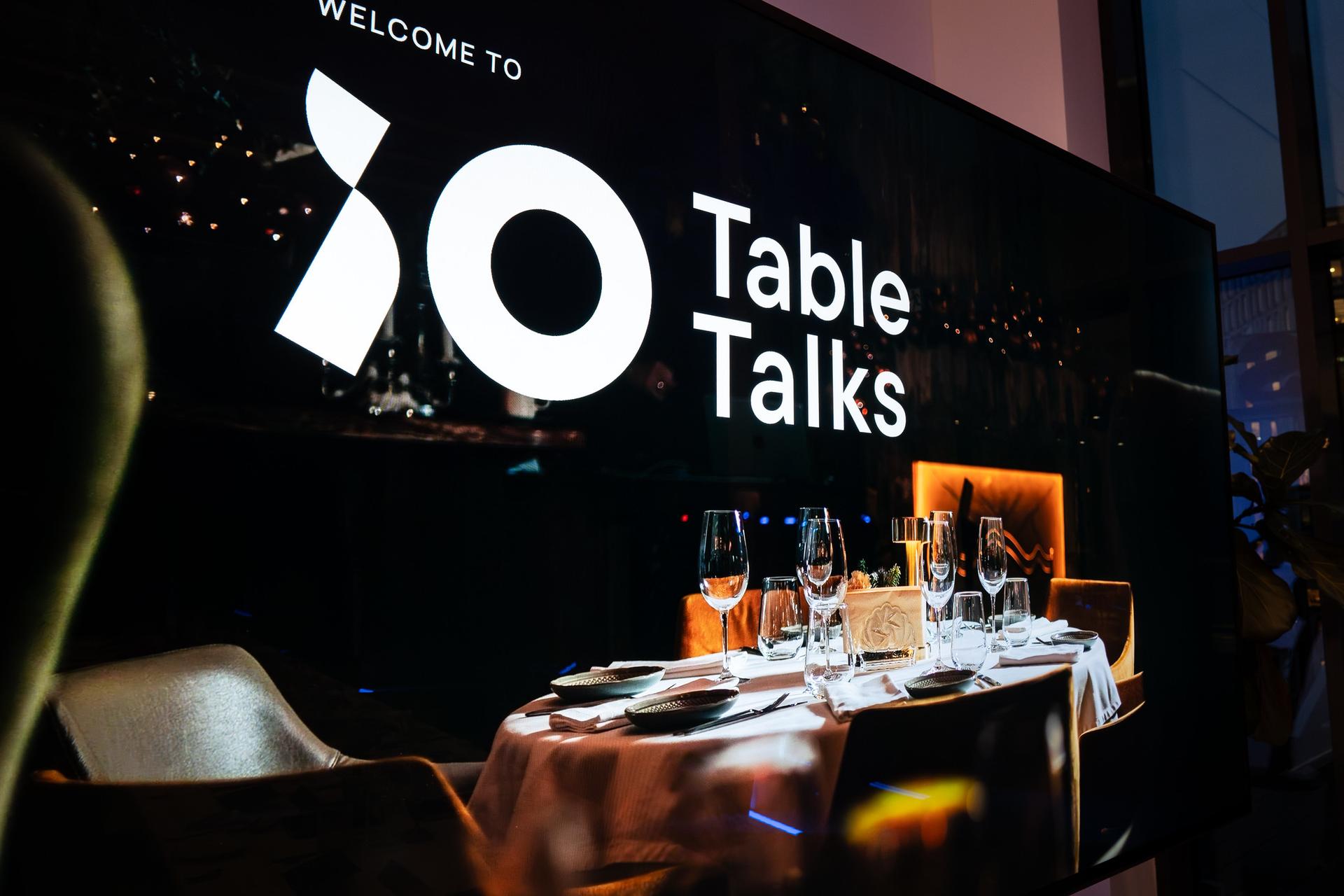 A screen displays "Welcome to Table Talks" with an image of a set dining table, featuring glasses and tableware under dim lighting.