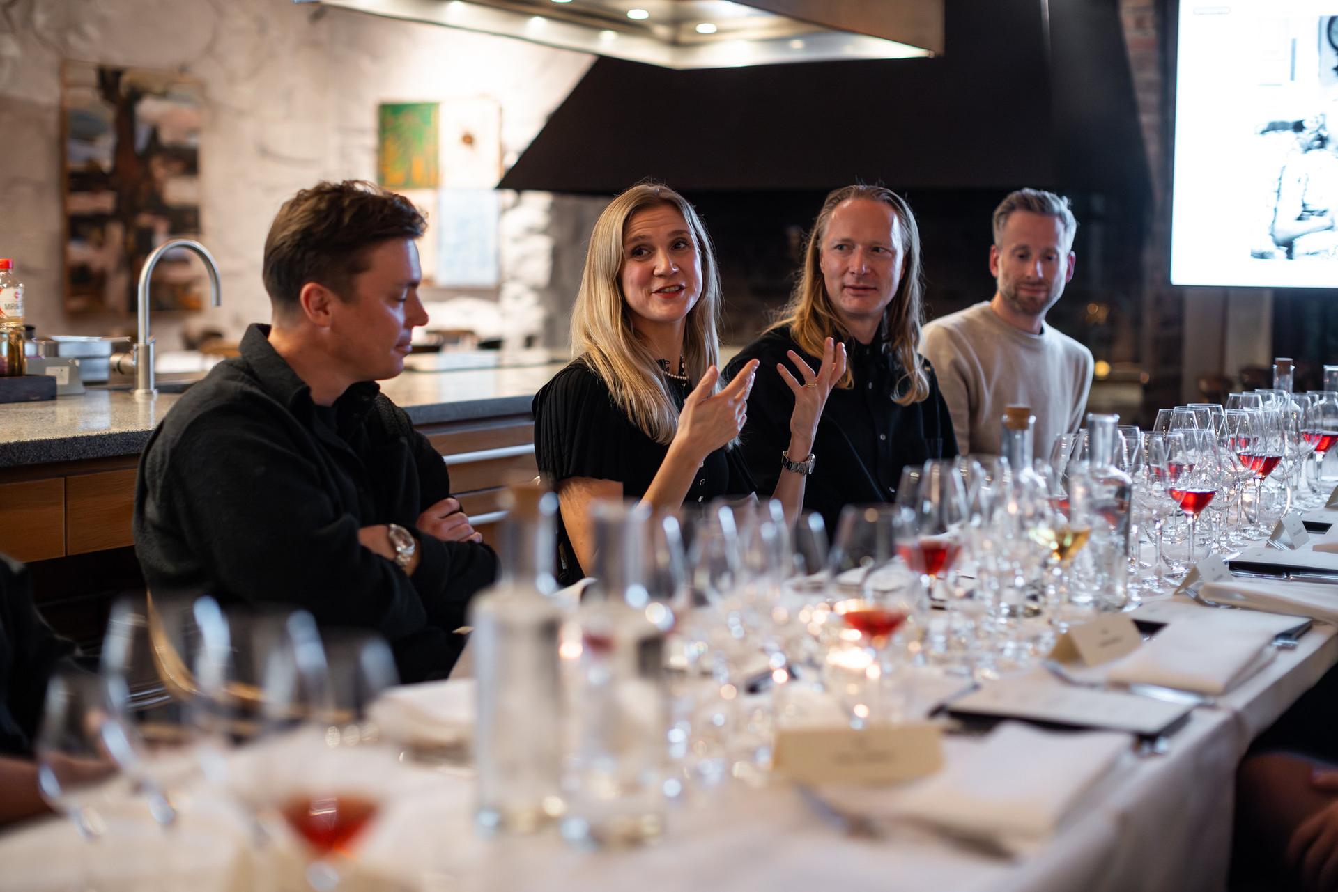 Four people sitting at a table covered with wine glasses, engaged in a discussion in a warmly lit room.