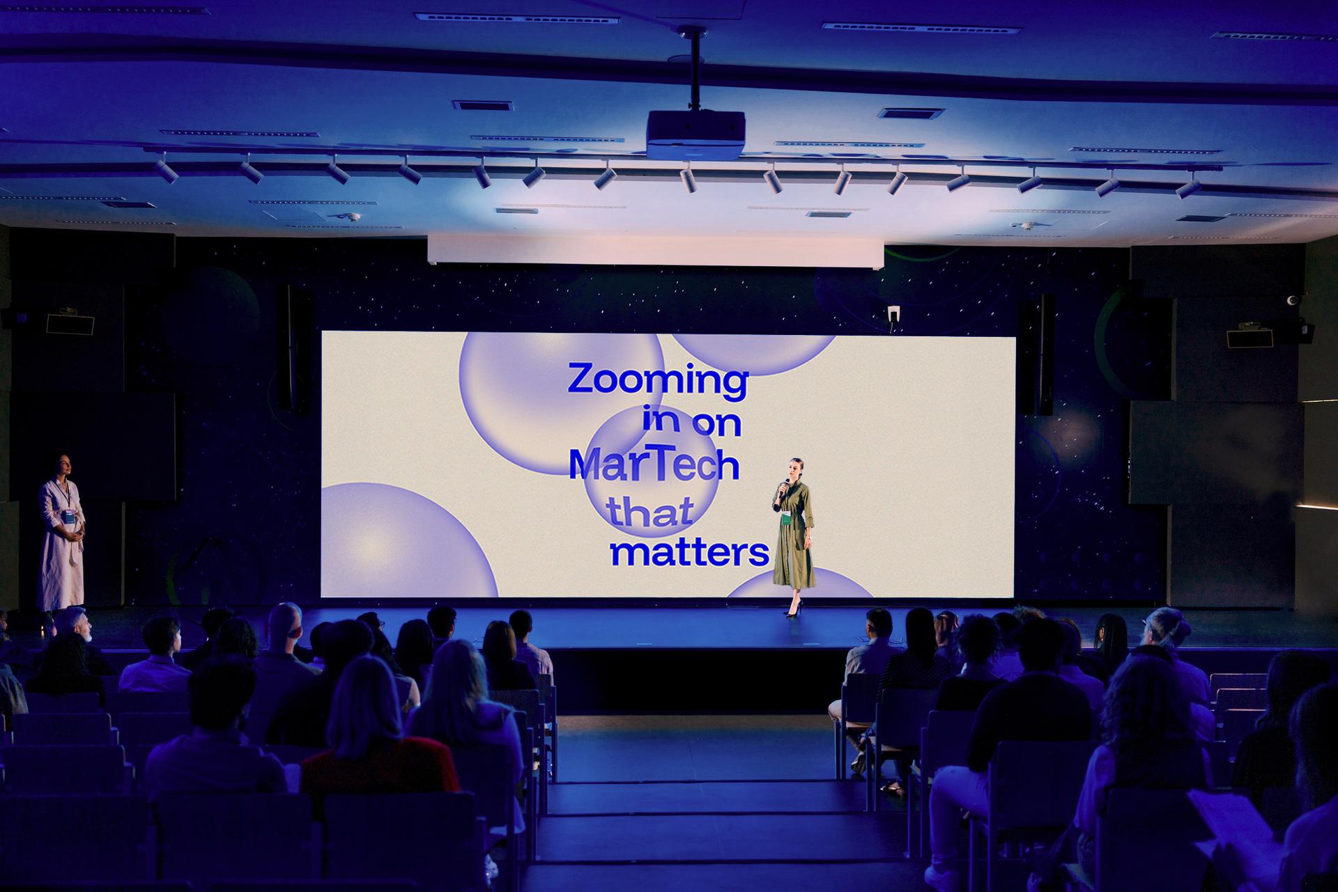 A speaker presents on stage with a large screen displaying "Zooming in on MarTech that matters" to an audience in a dimly lit auditorium.