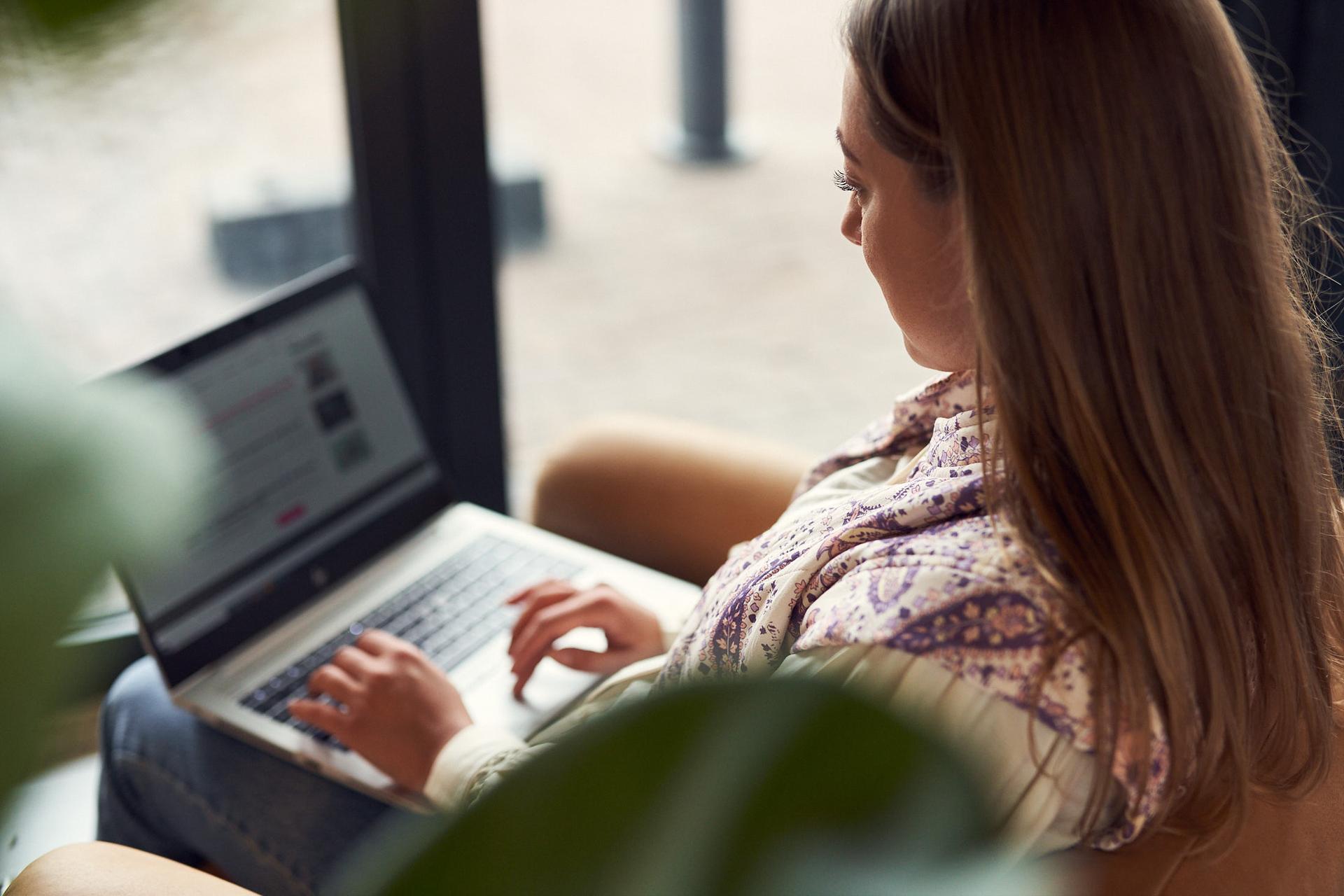 Woman with long hair using a laptop, sitting by a large window, partially obscured by plant leaves in the foreground.