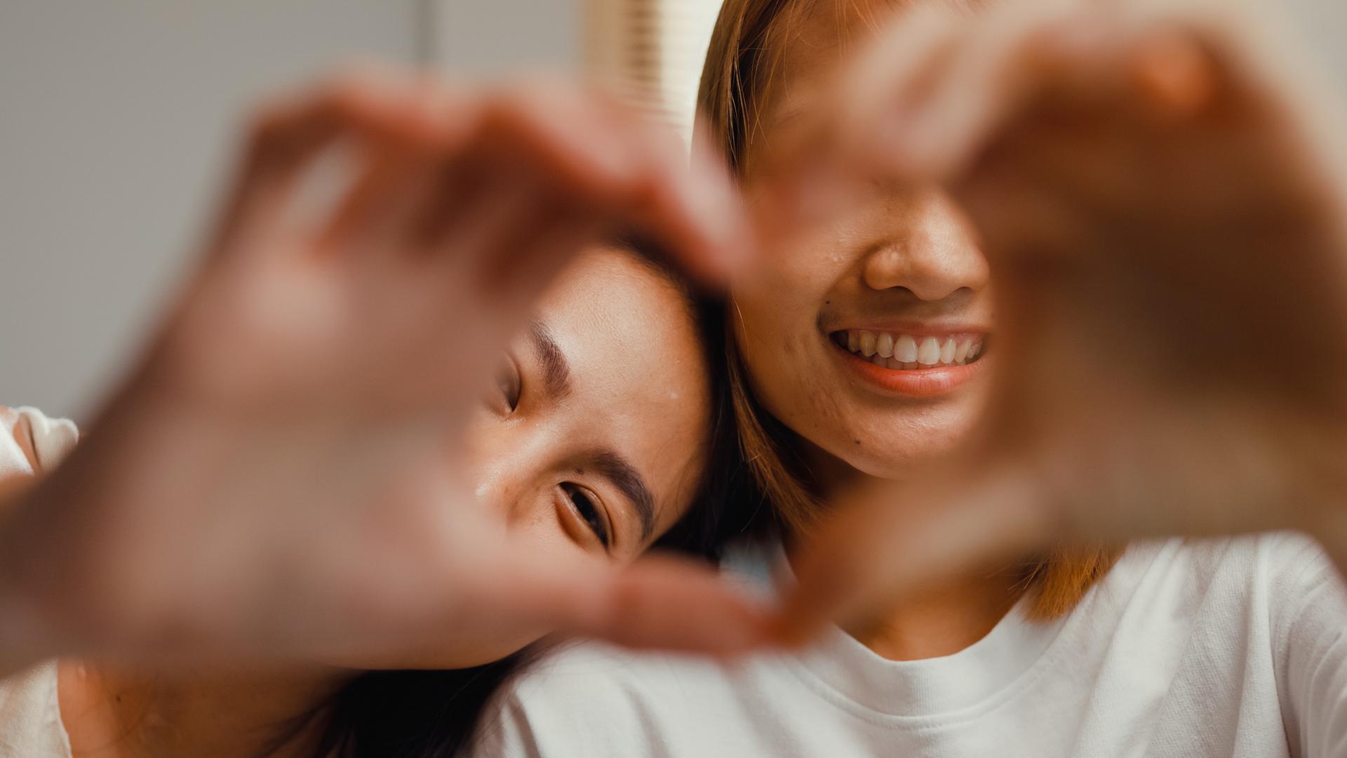 Two people smiling and forming a heart shape with their hands in the foreground, creating a joyful and friendly atmosphere.