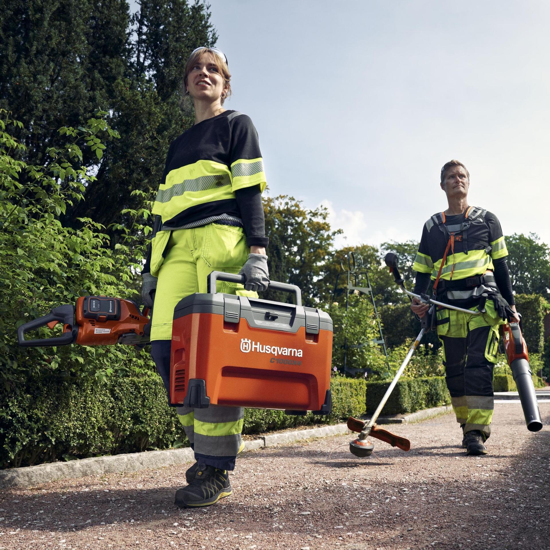 Two workers in high-visibility gear carry Husqvarna tools, including a chainsaw and a leaf blower, while walking on a garden path.