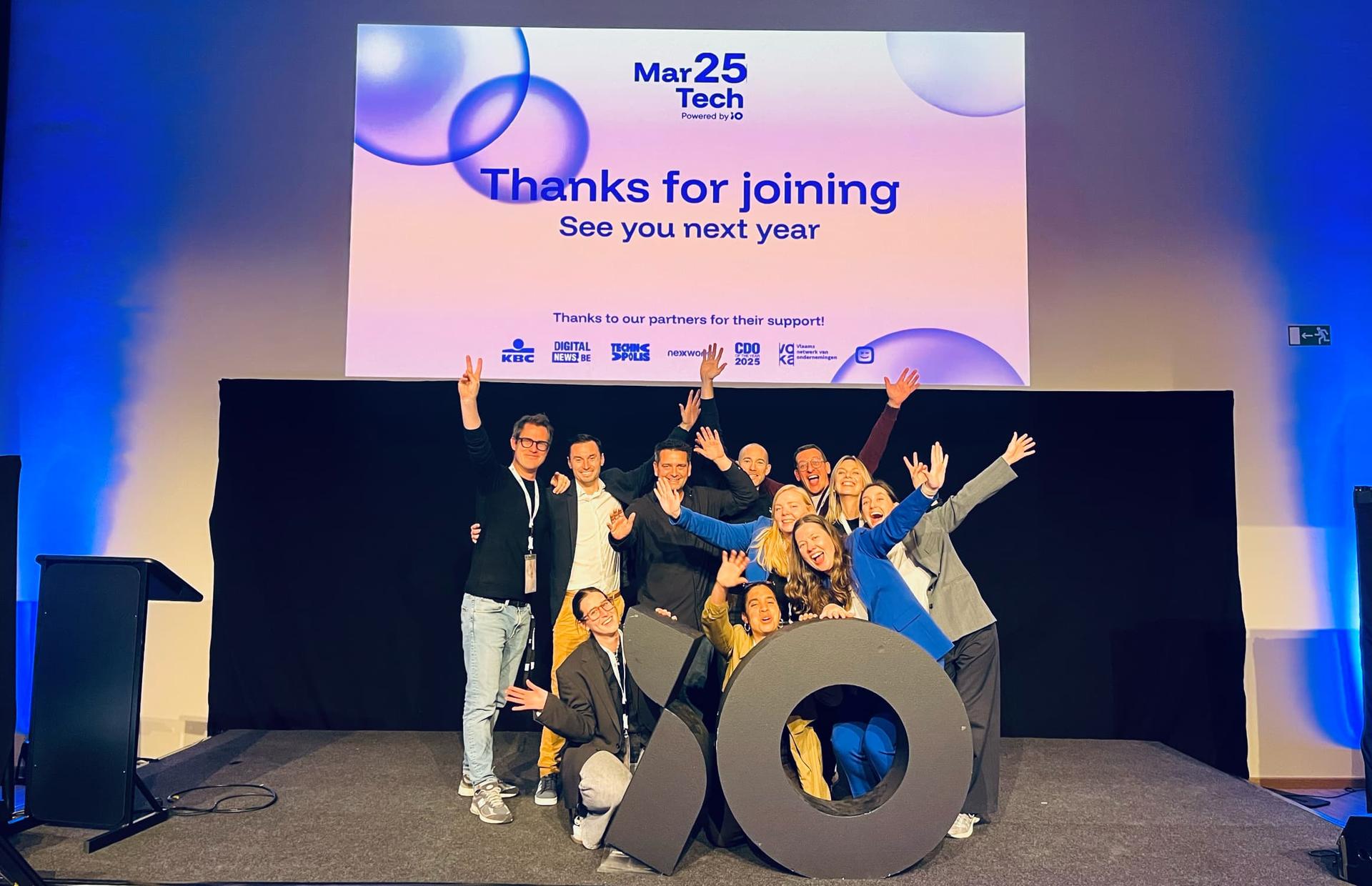 A group of people posing energetically on stage in front of a large screen displaying "Thanks for joining. See you next year" at a tech event.