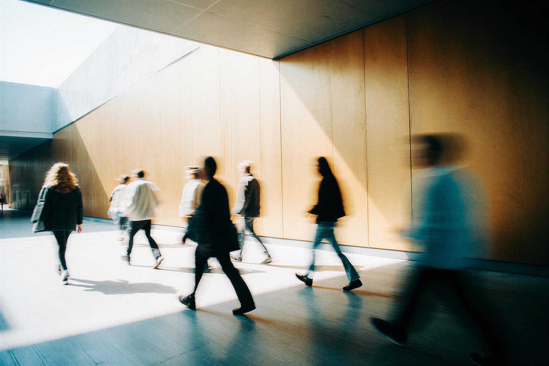 Blurred figures walking in a sunlit hallway with wooden walls, casting soft shadows on the floor.