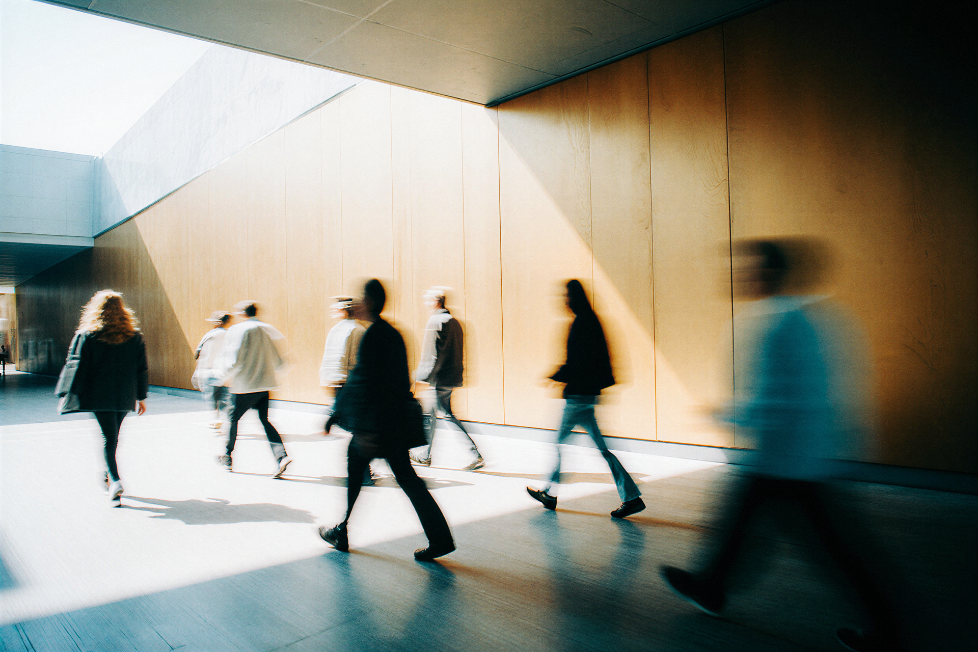 Blurred figures walking in a sunlit hallway with wooden walls, creating a sense of motion and bustling activity.