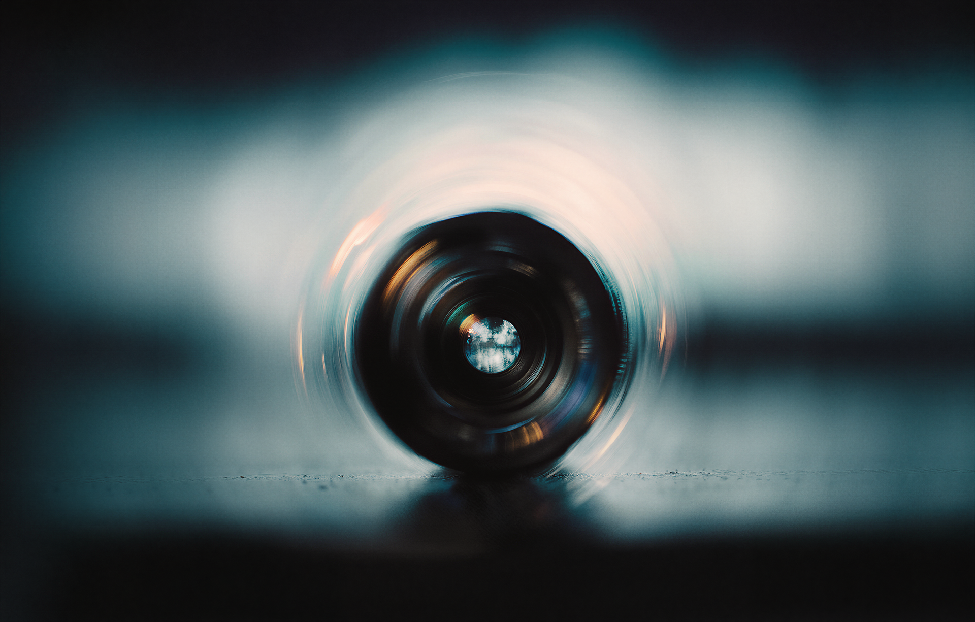 Blurred close-up of a camera lens on a reflective surface, surrounded by soft, swirling glow with blue and orange hues.