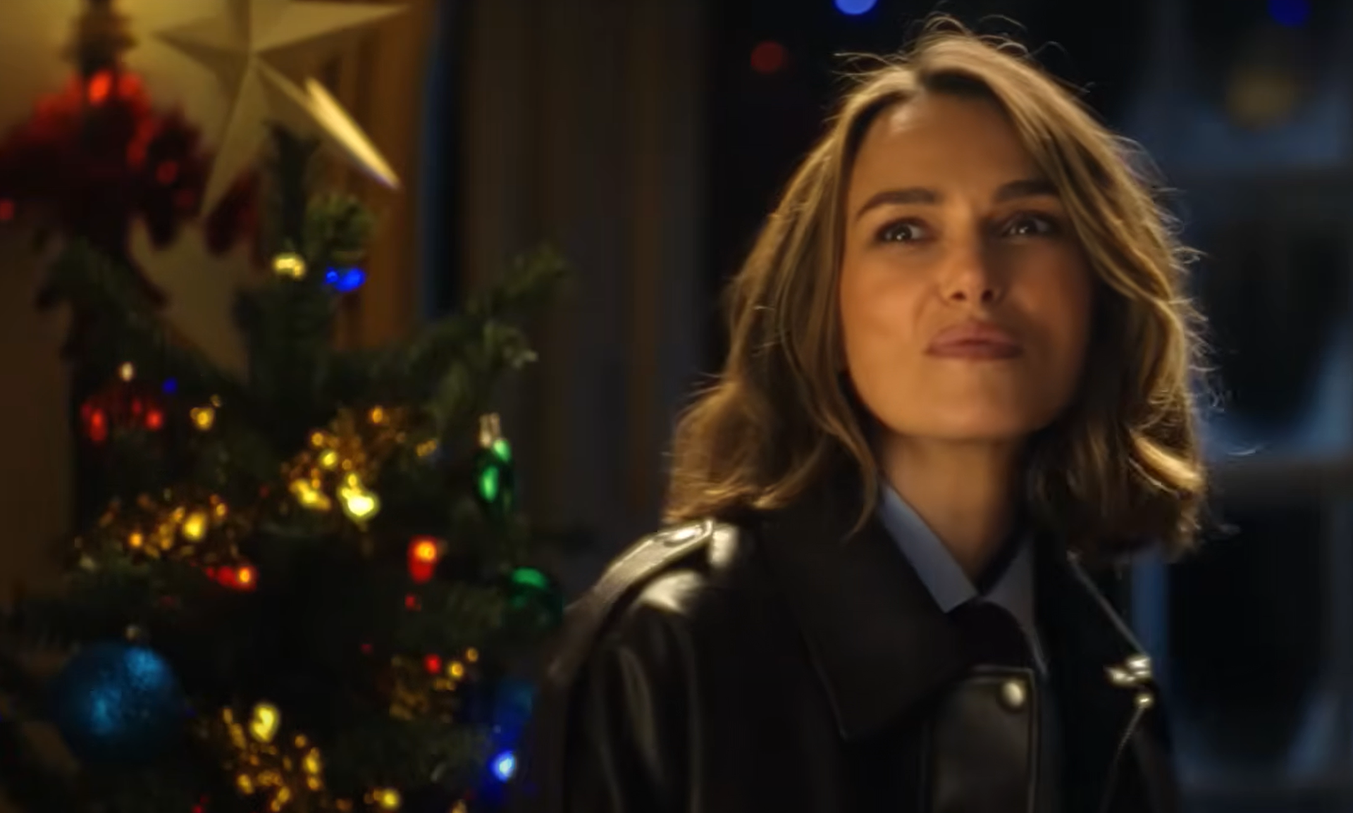 Keira Knightley in a leather jacket smiles near a decorated Christmas tree with colorful lights and ornaments.