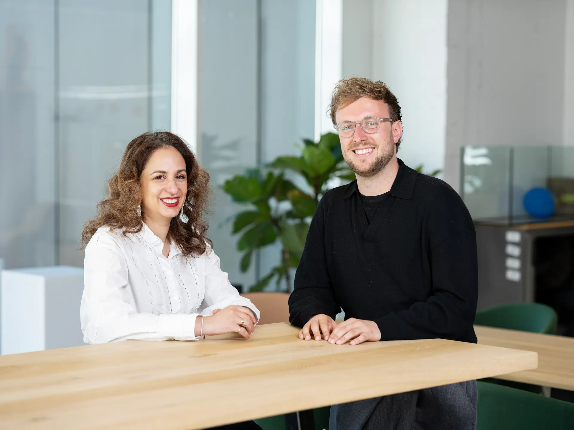 Two people smiling while sitting at a wooden table in a modern office setting with a plant in the background.