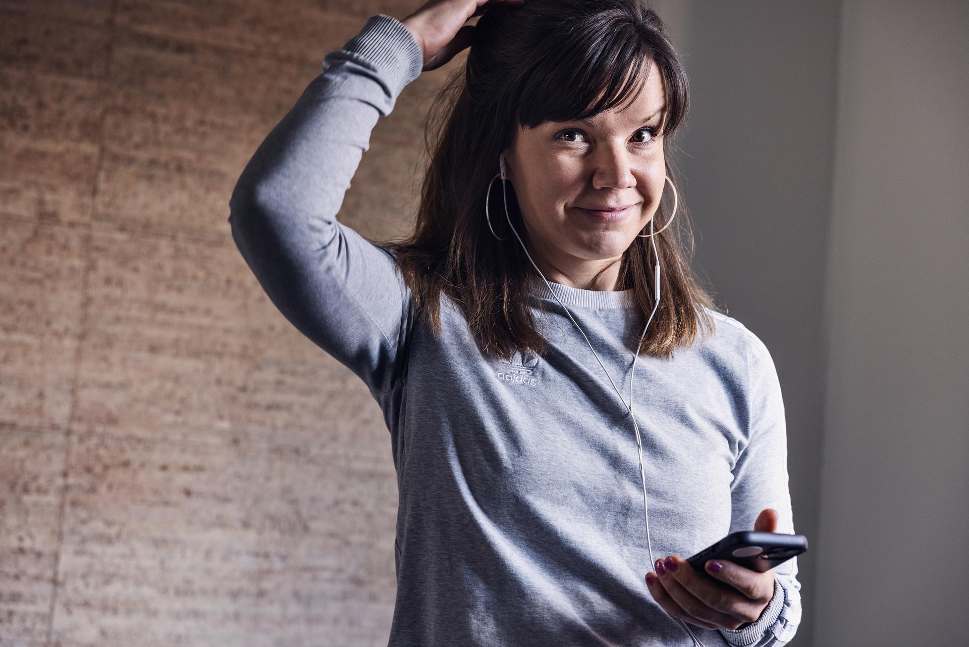 Woman in a gray sweater smiles while holding a smartphone and adjusting her hair, wearing earphones.