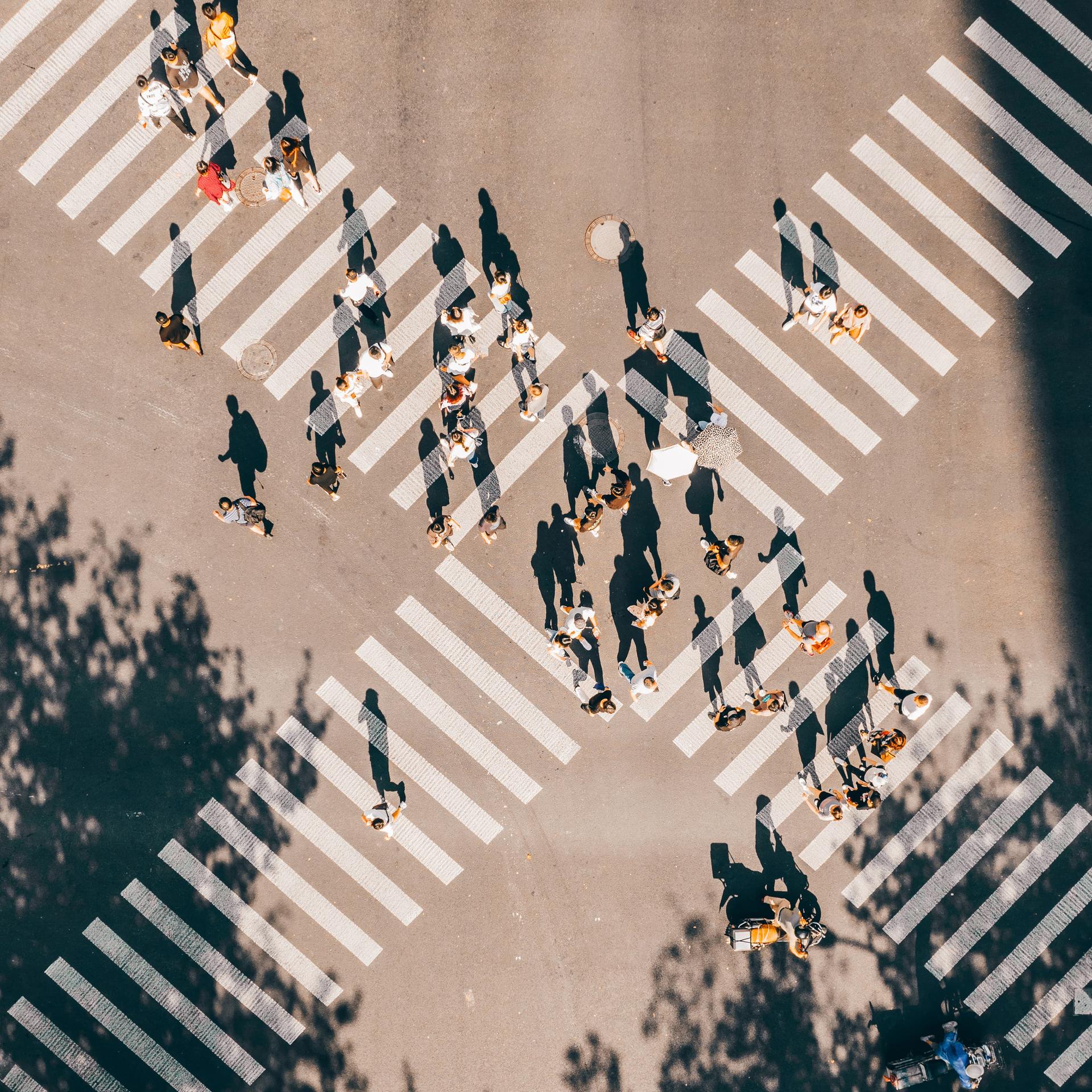 Aerial view of a busy crosswalk with people crossing in different directions, casting long shadows on the sunlit street.