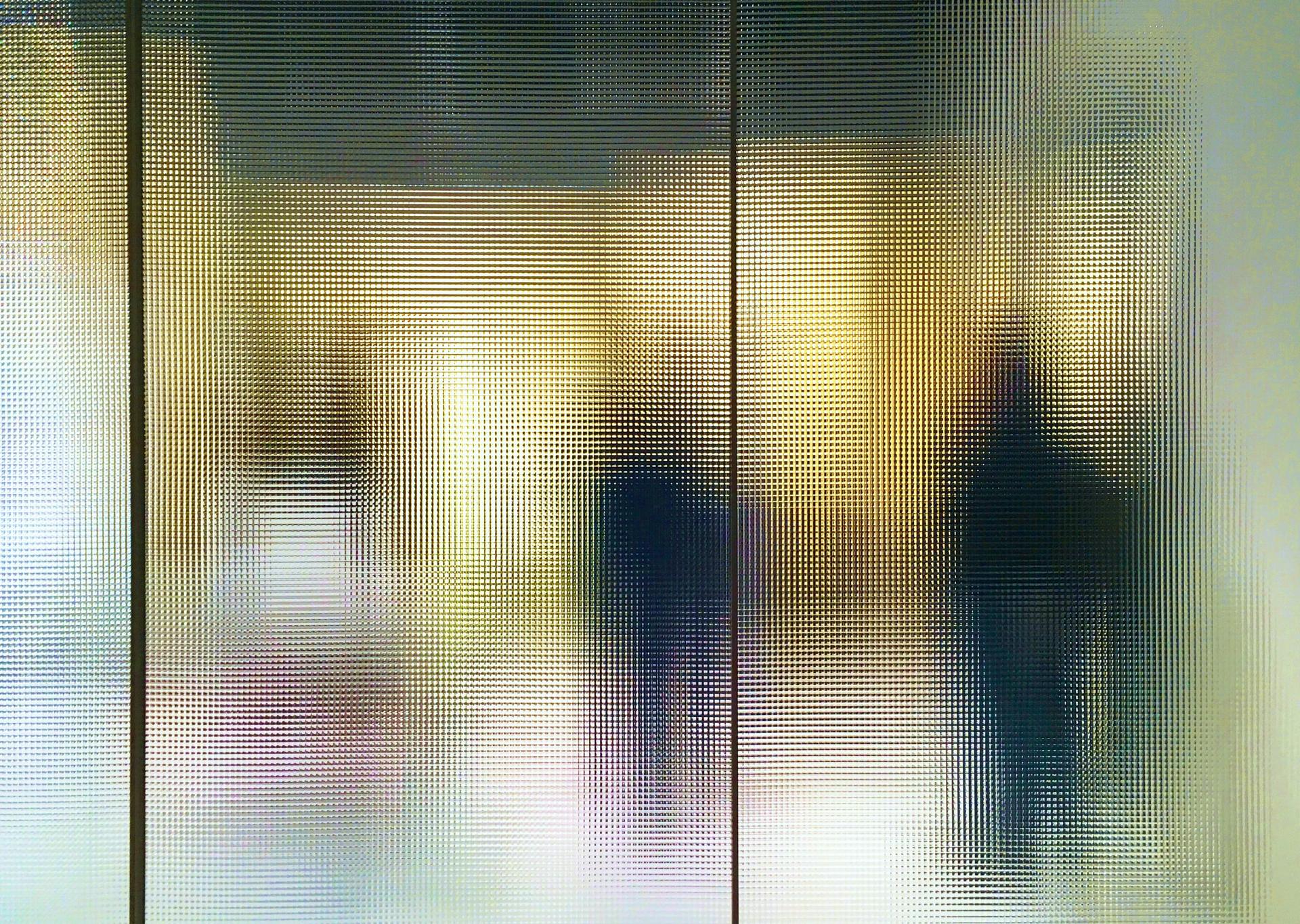 Abstract image of blurred silhouettes behind a textured glass surface, creating a soft, distorted view with vertical lines.