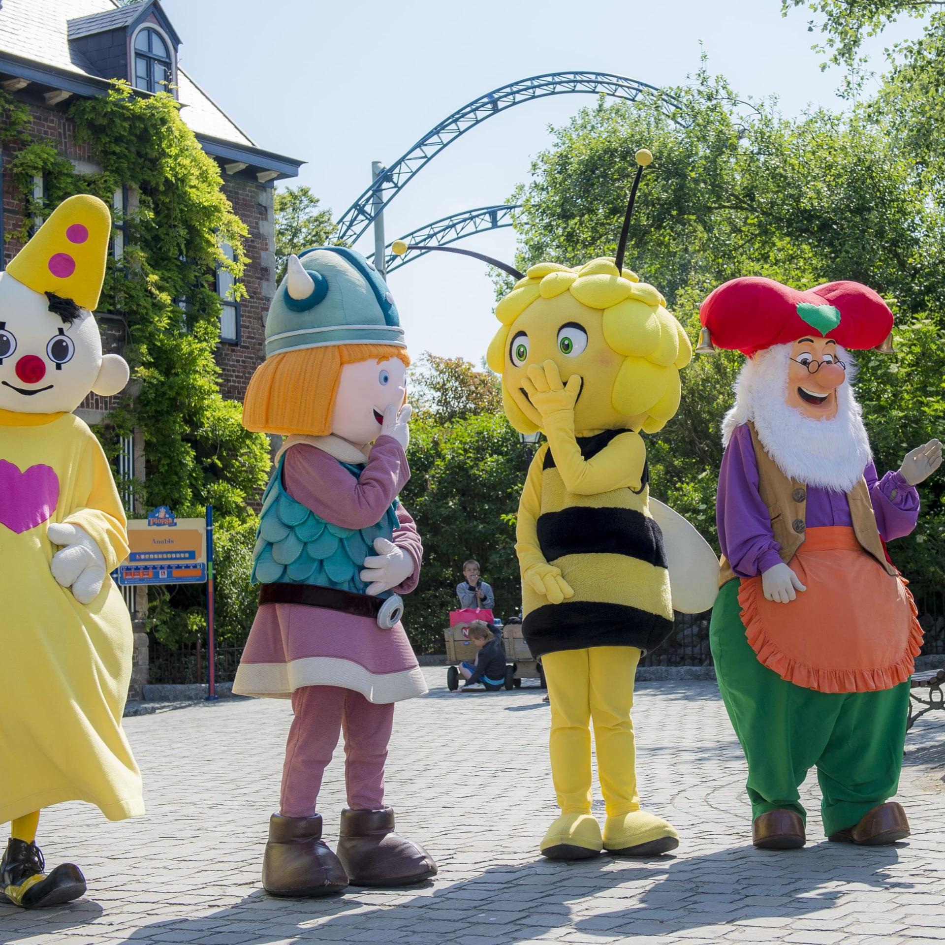 Four people in colorful mascot costumes, including a clown, Viking, bee, and gnome, stand on a paved path with trees and a ride in the background.
