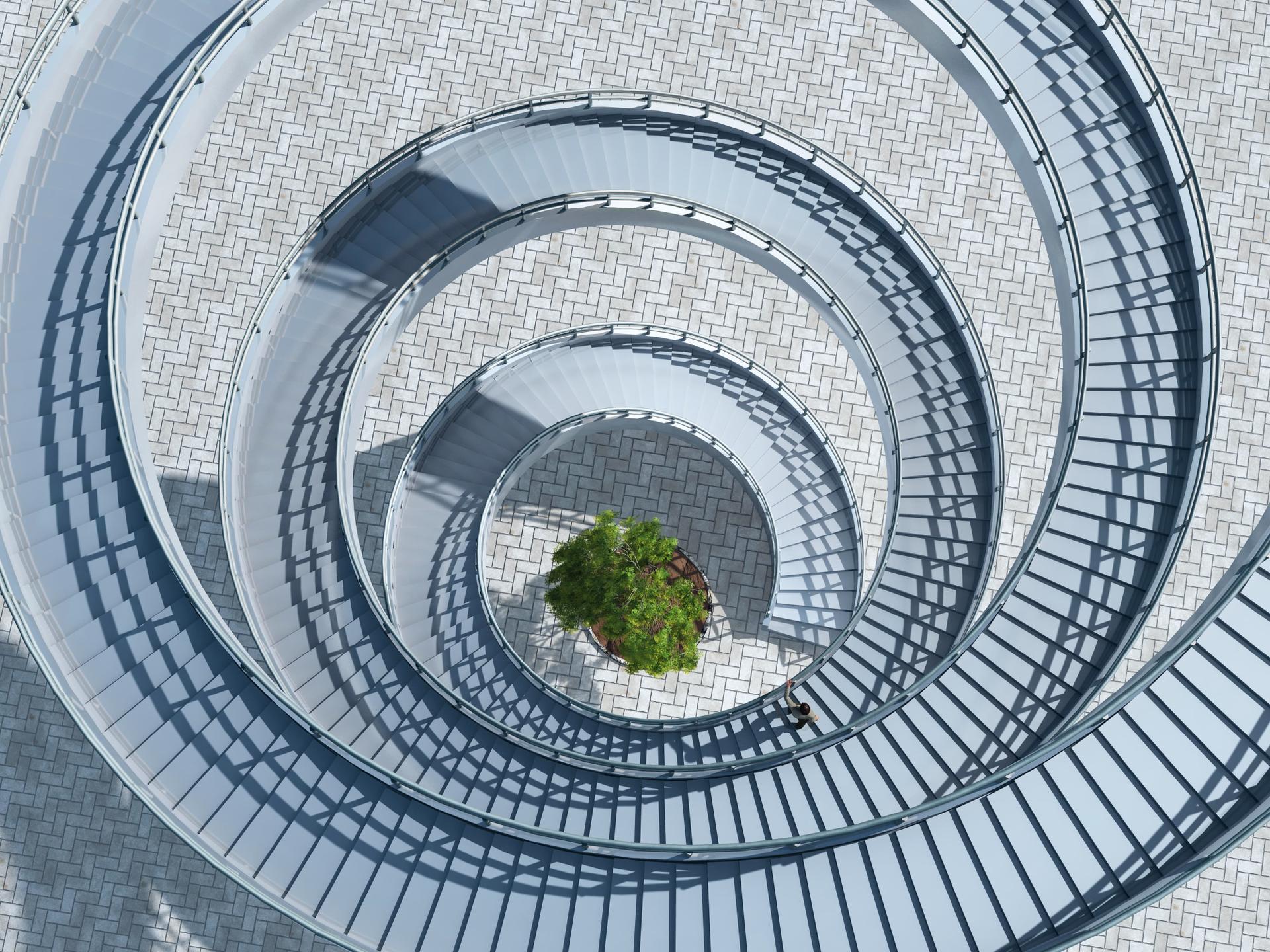Aerial view of a winding spiral staircase with metal railings, leading down to a small tree on a patterned brick surface.