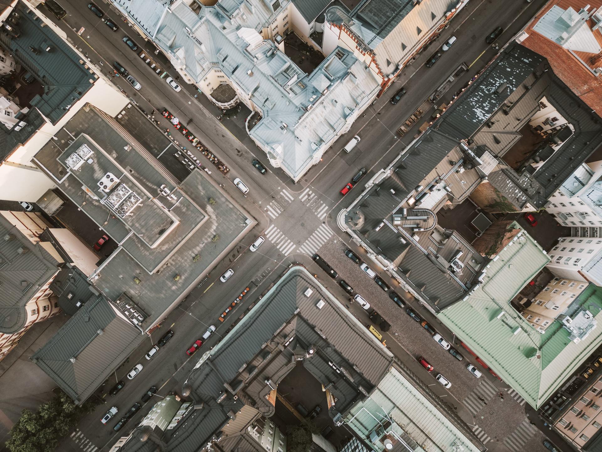 Aerial view of an urban intersection surrounded by buildings, with cars parked along the streets and pedestrians crossing.