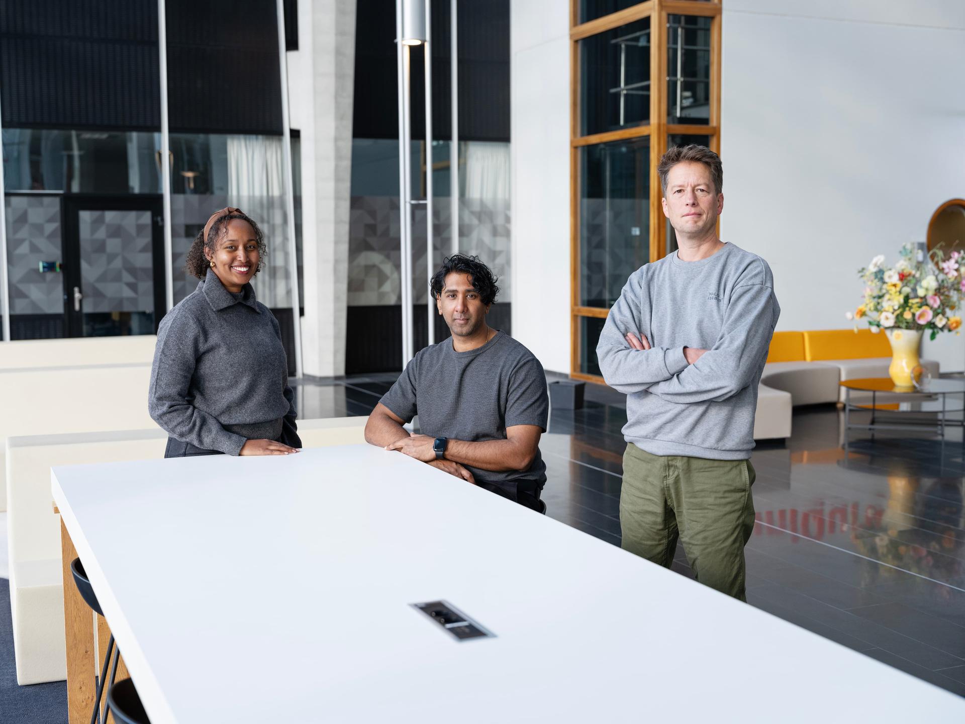 Three people in casual attire stand and sit around a white table in a modern, spacious room with large windows and a floral arrangement.