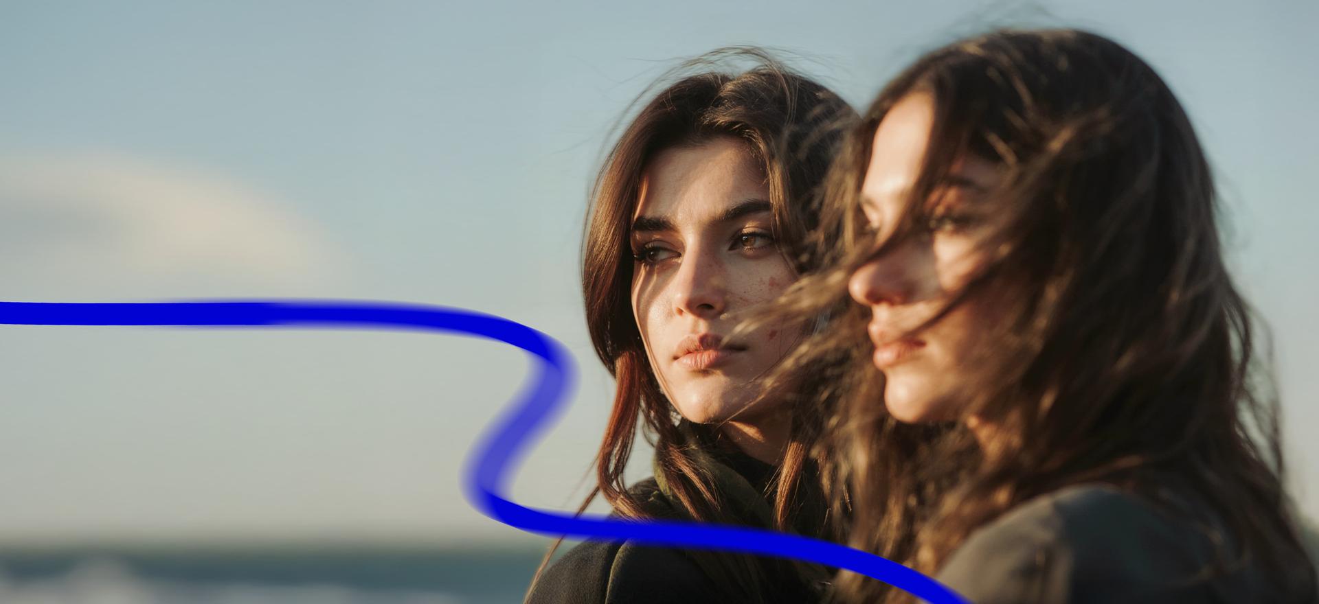 Two women with long hair stand close together outdoors, gazing in the same direction. A blue wavy line crosses the image.