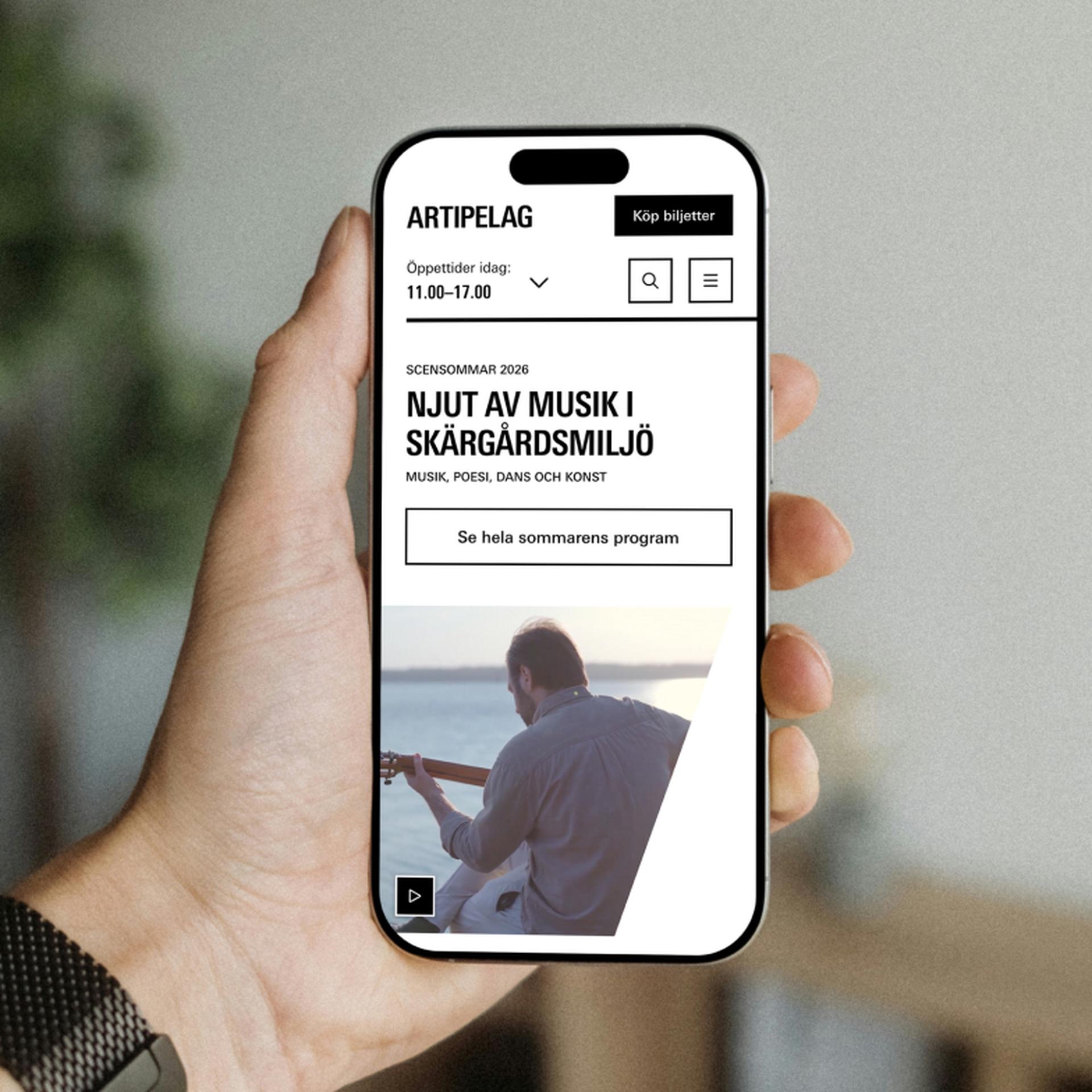 Hand holding a smartphone displaying an event page for "Artipelag" featuring music, poetry, dance, and art in a scenic archipelago setting.