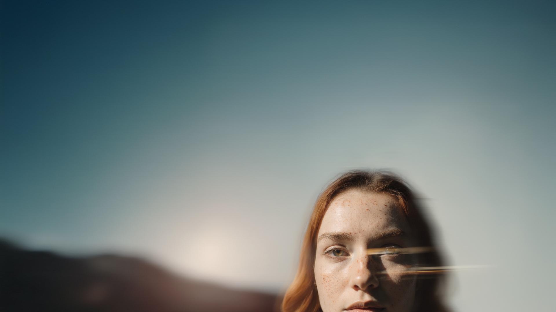 A woman partially visible against a bright blue sky with sunlight reflecting on the camera lens.