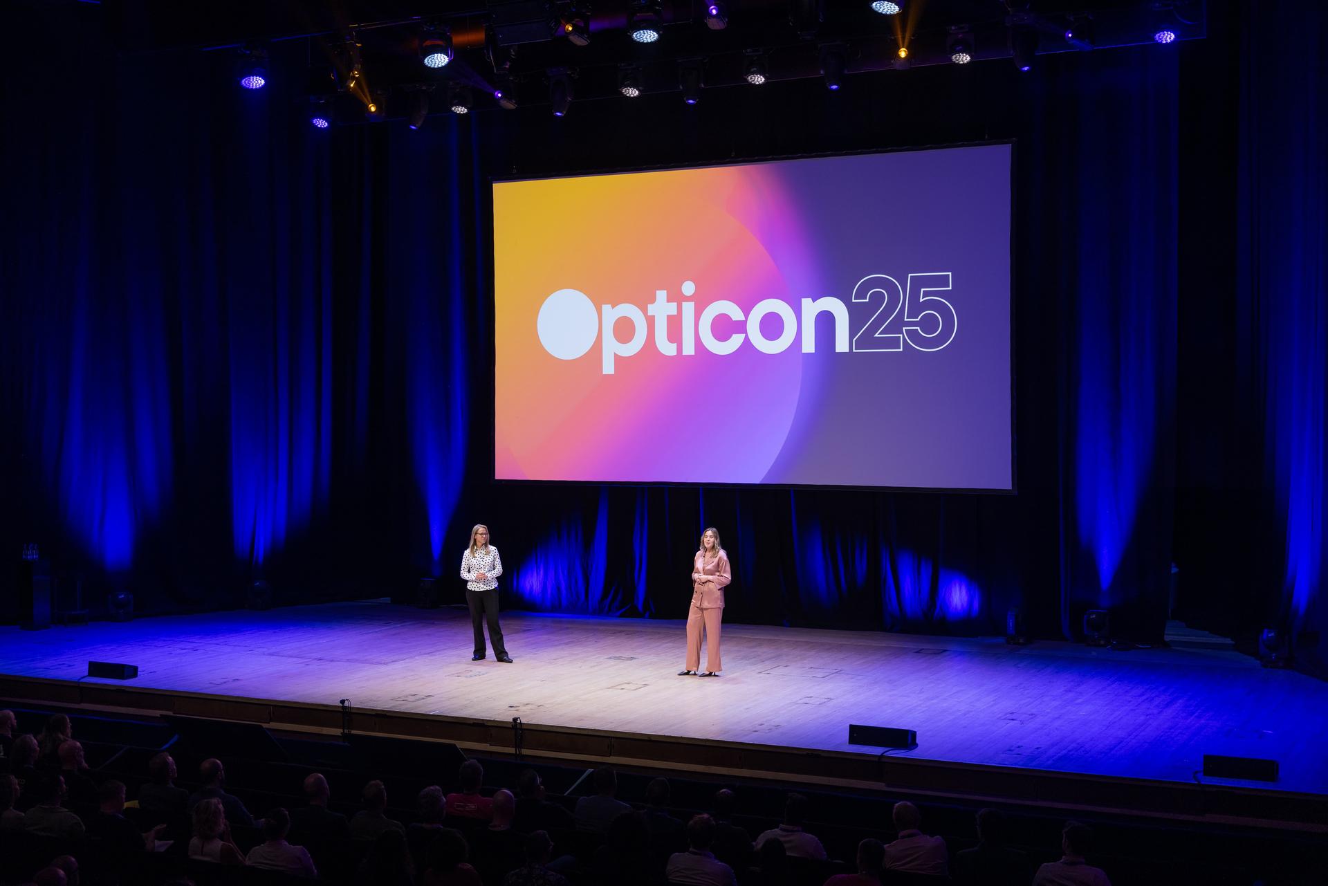 Two presenters on stage at Opticon 25 conference, with a large screen displaying the event logo, in a dimly lit auditorium.