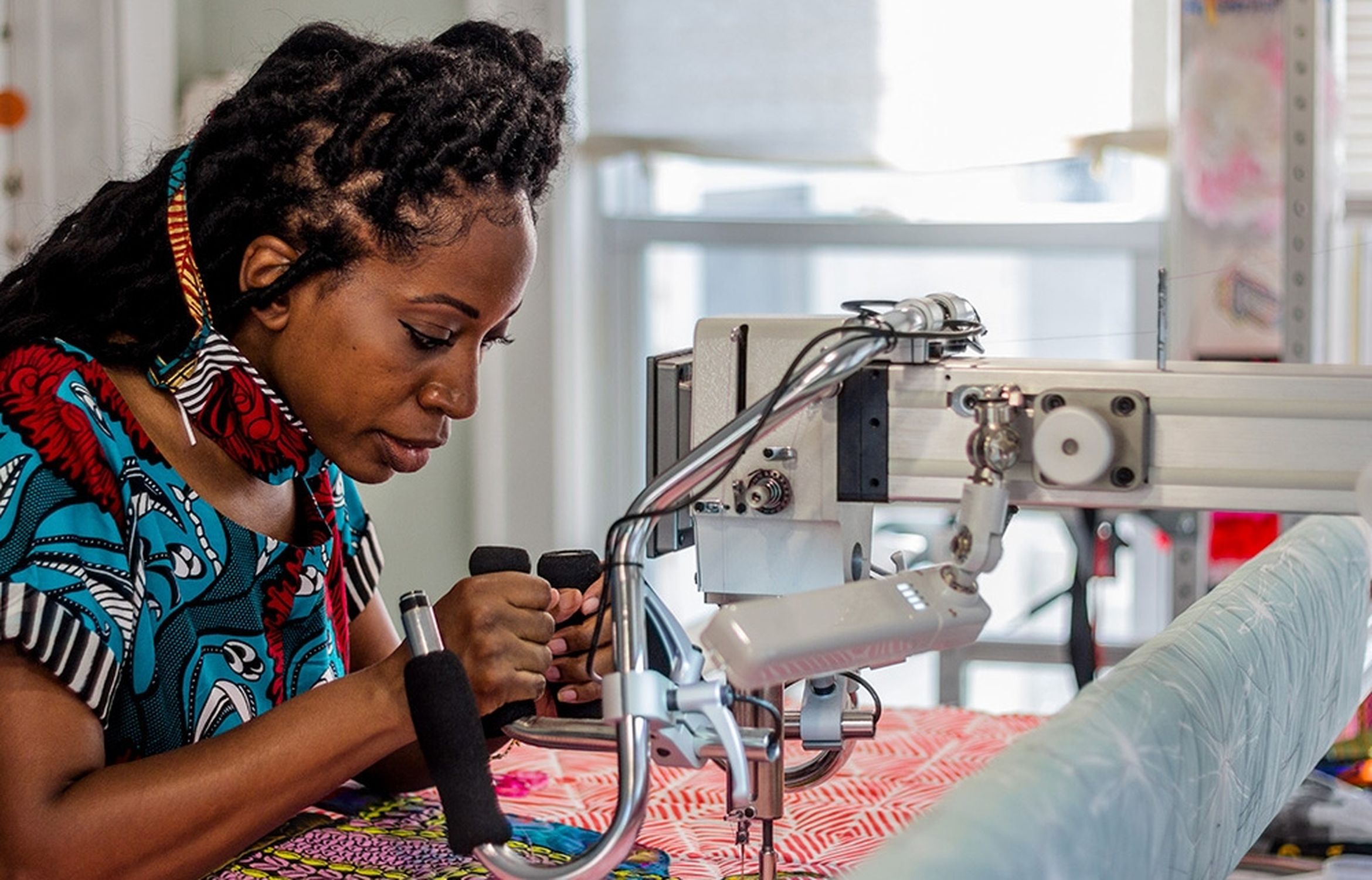 Artist Bisa Butler intensely focuses on operating a quilting machine, meticulously working on a colorful fabric with intricate patterns in a bright room.