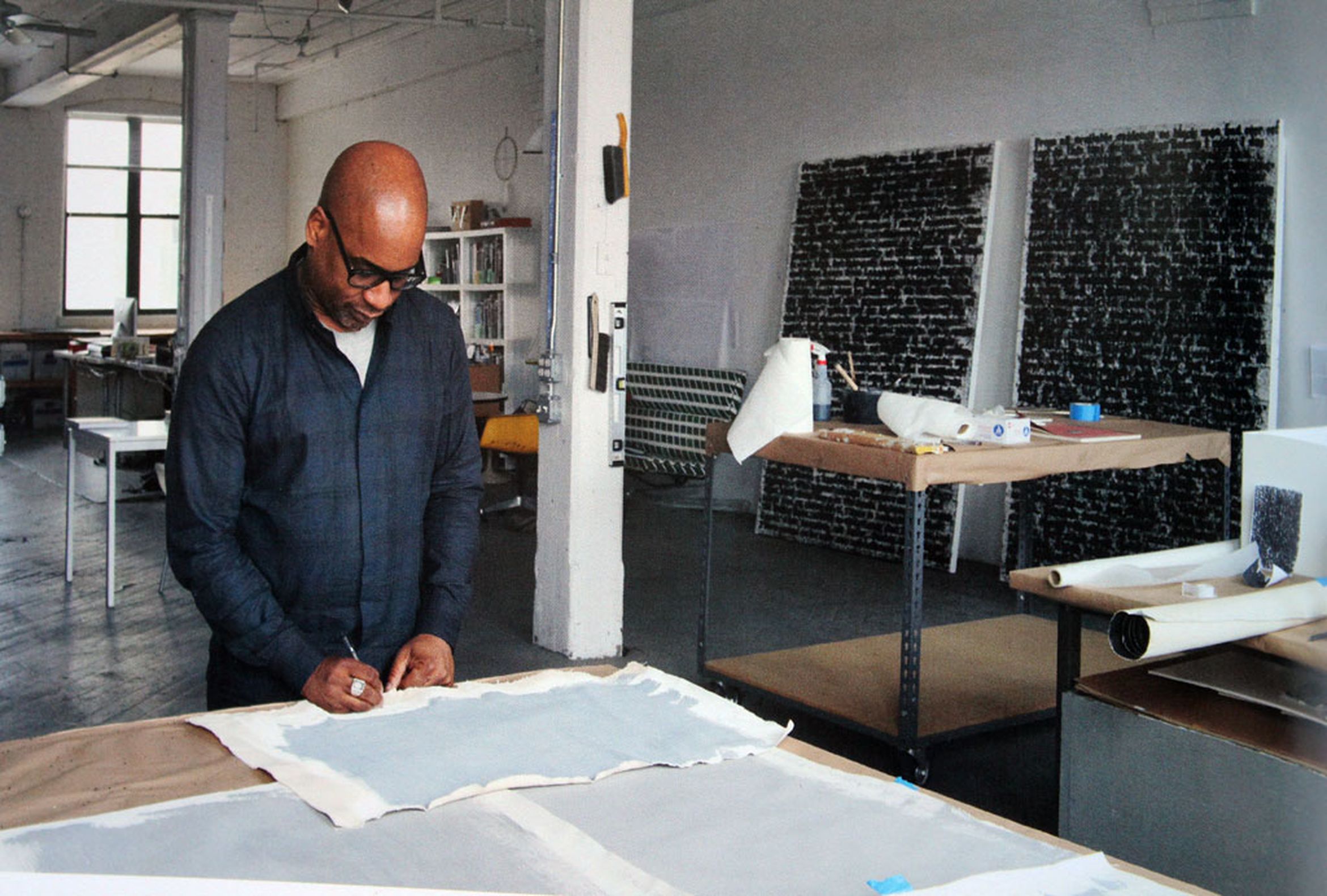 Artist (Glenn Ligon) in a black shirt works on a canvas in a spacious, light-filled studio with art supplies and completed artworks in the background.