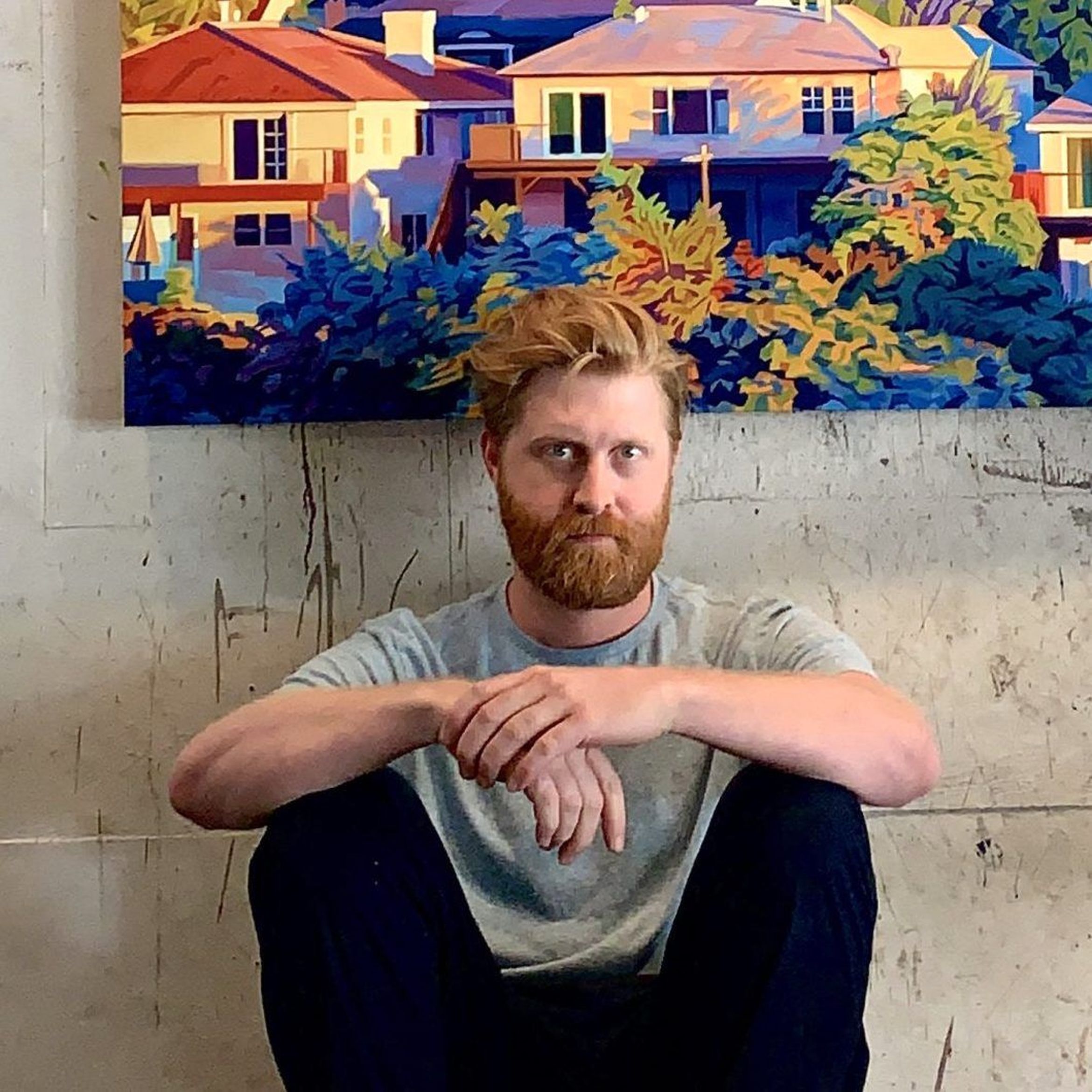 A person with a beard and casual attire sits on the floor in front of a colorful painting of houses and foliage.