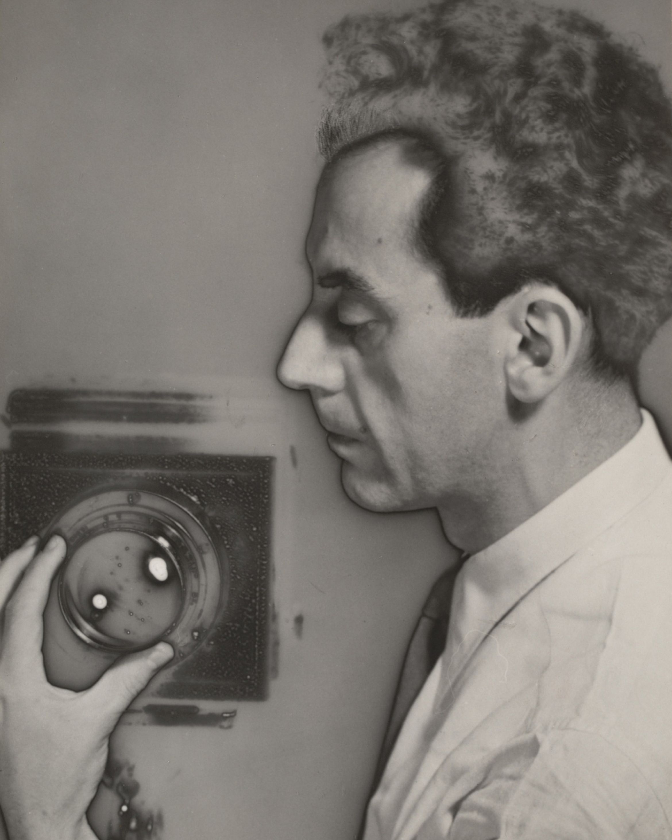 Black and white profile of a man with curly hair, examining a glass object against a textured surface.