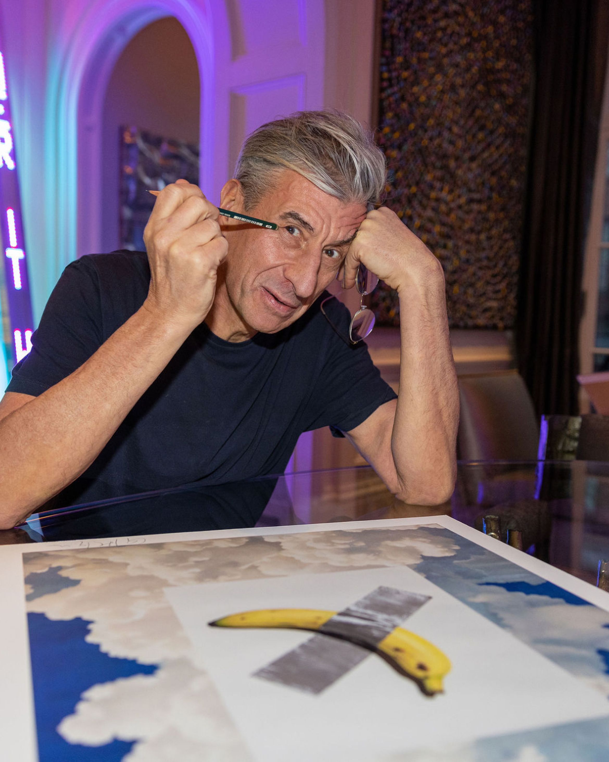 Man playfully holds glasses near a surreal banana print on a table. Neon light and textured wall in background.