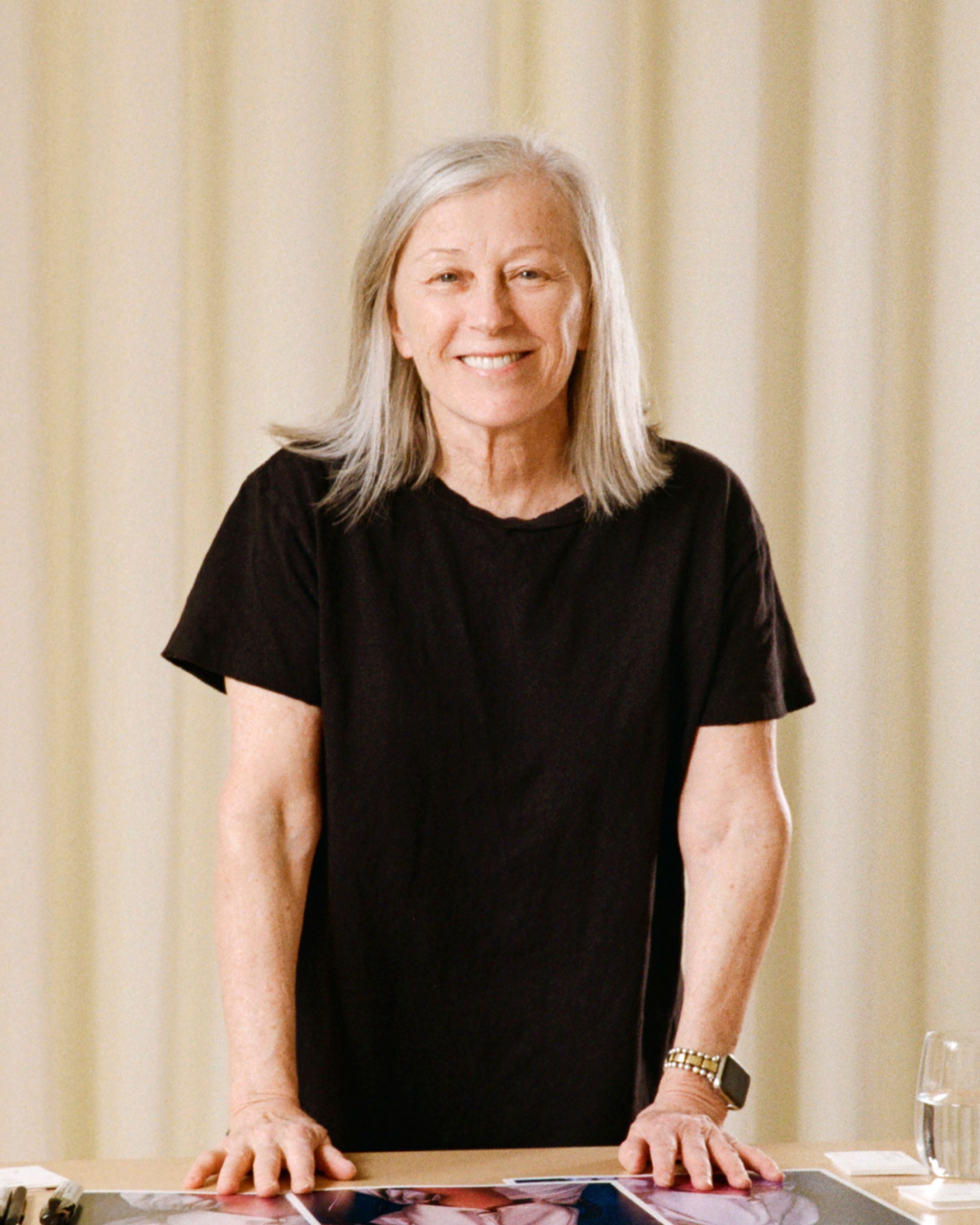 Person in a black shirt standing at a table, smiling, with a glass of water nearby and a light curtain in the background.
