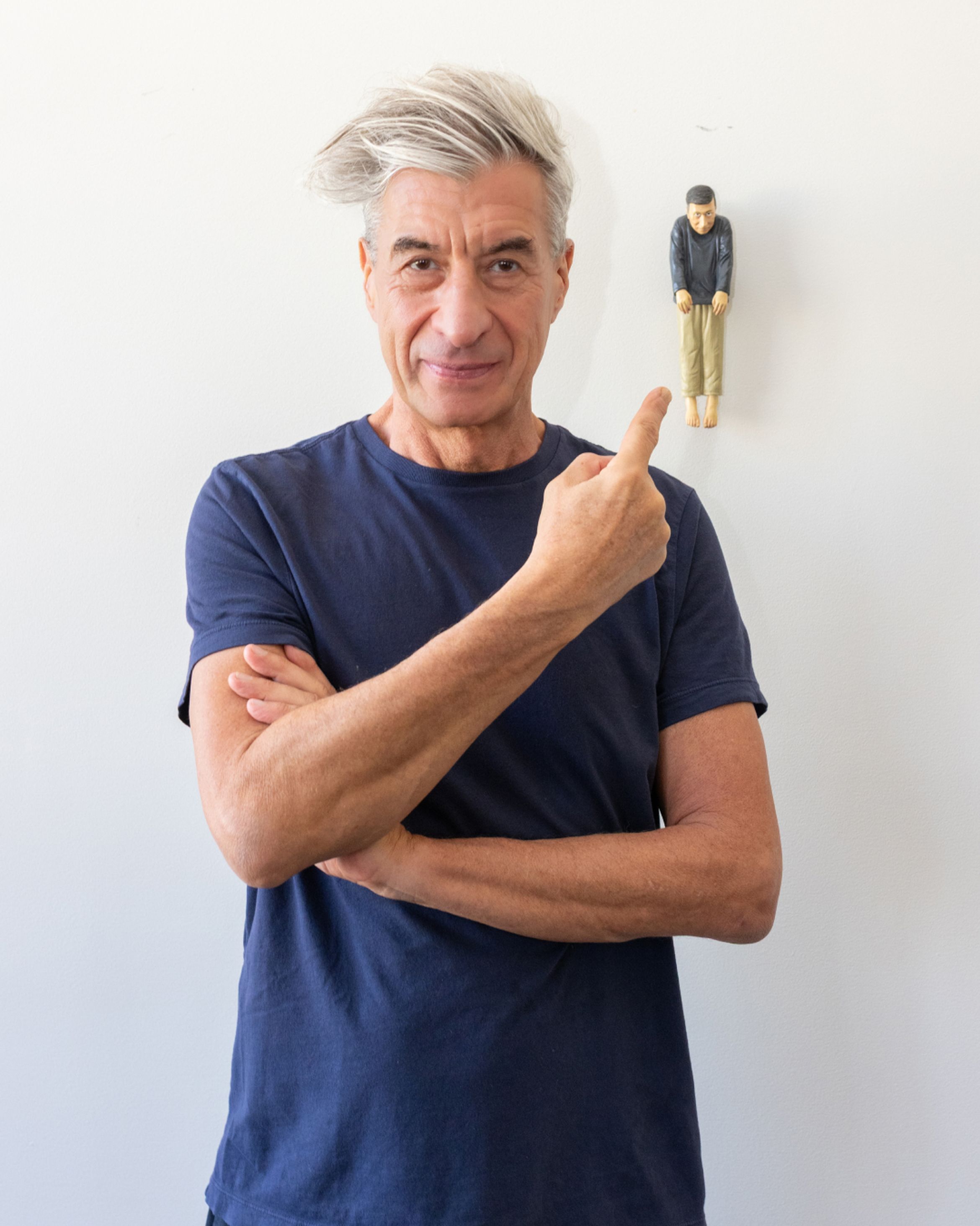 Man in a blue shirt with gray hair points at a small figurine on a white wall, smiling with arms crossed.