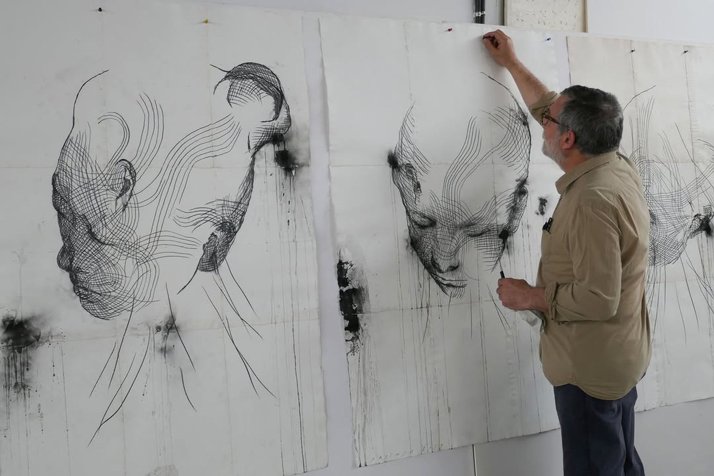 Artist drawing intricate line art of faces on large paper sheets in a studio.