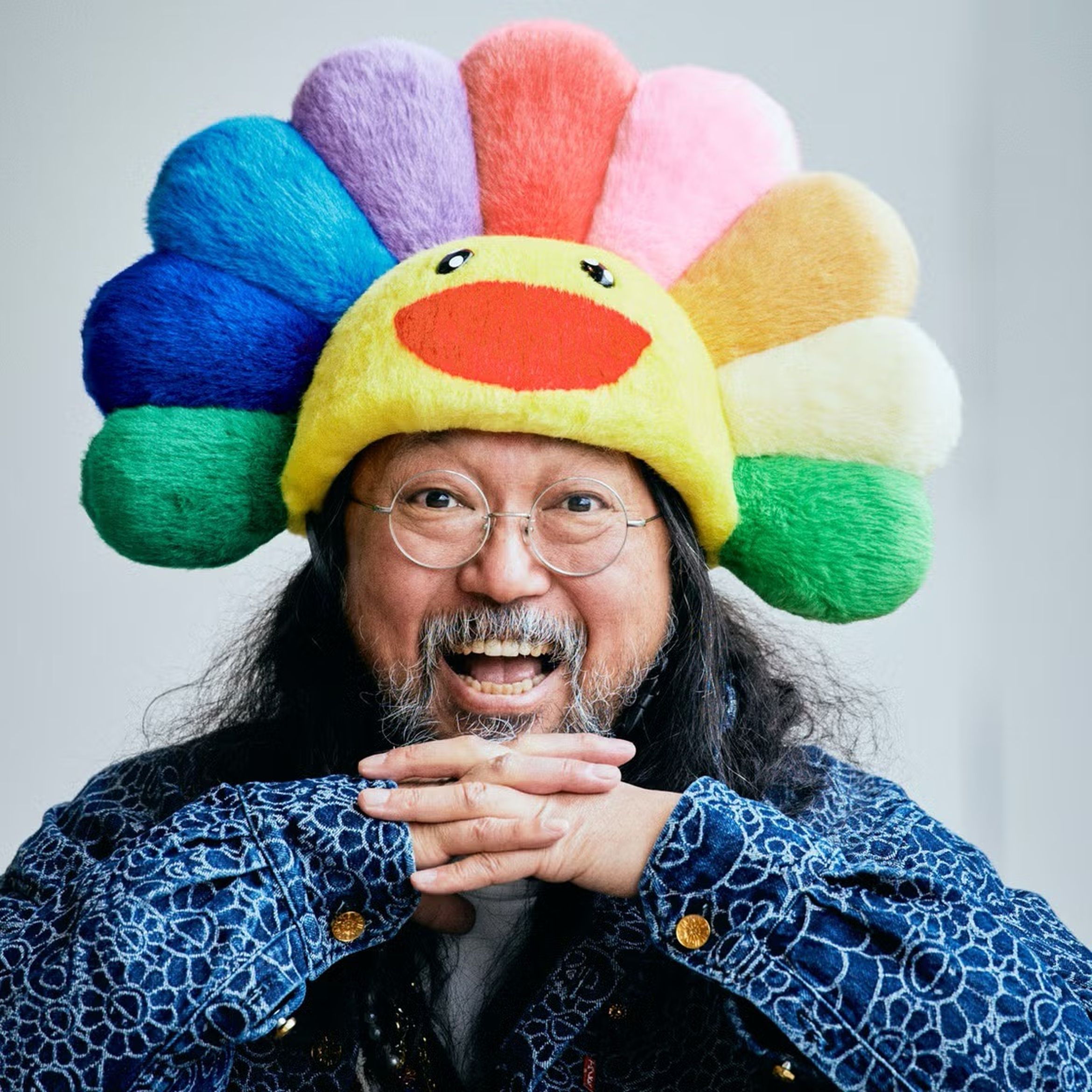 A person with long hair and glasses smiles, wearing a colorful, plush hat shaped like a flower with a face, against a plain background.