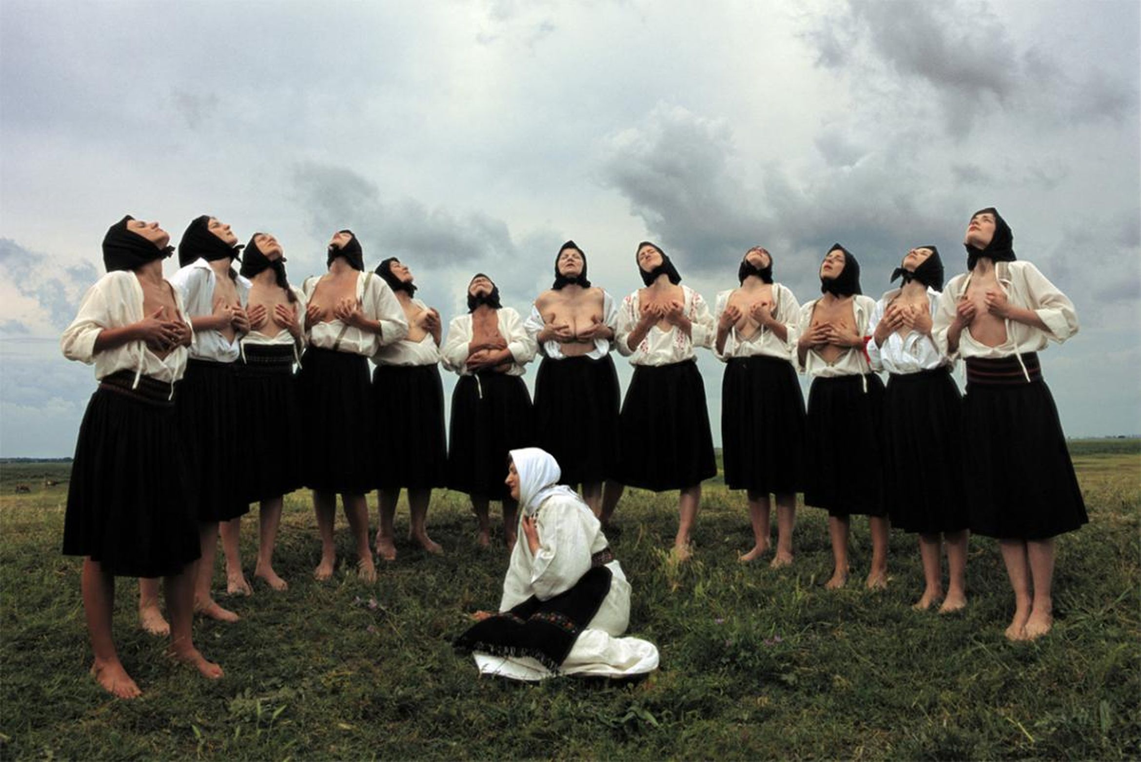 Women in traditional attire with veils, standing in a grassy field, looking upward with open shirts, while one kneels in front.