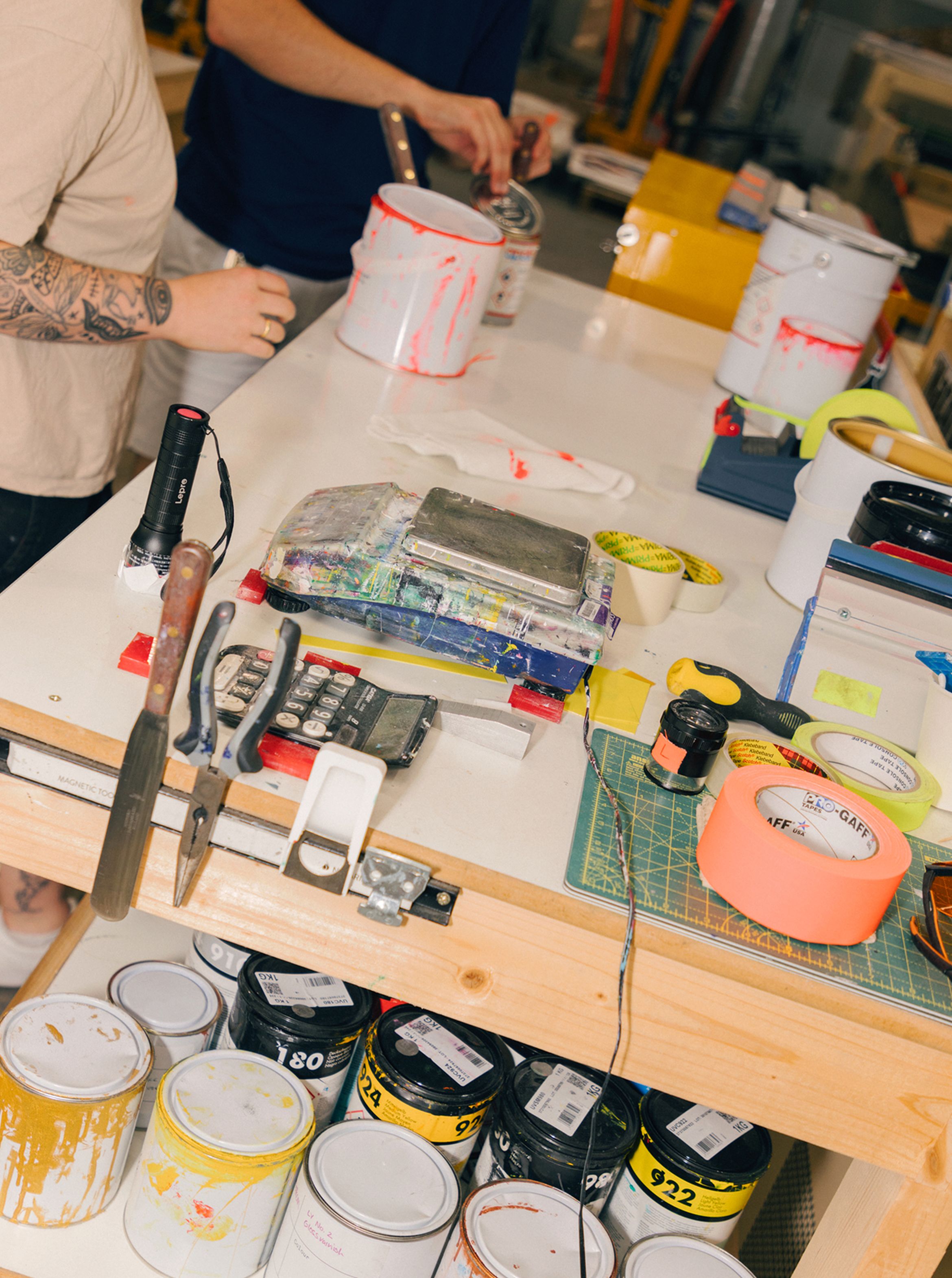 A cluttered workbench with paint cans, brushes, tape, and tools. Two people are working in the background.