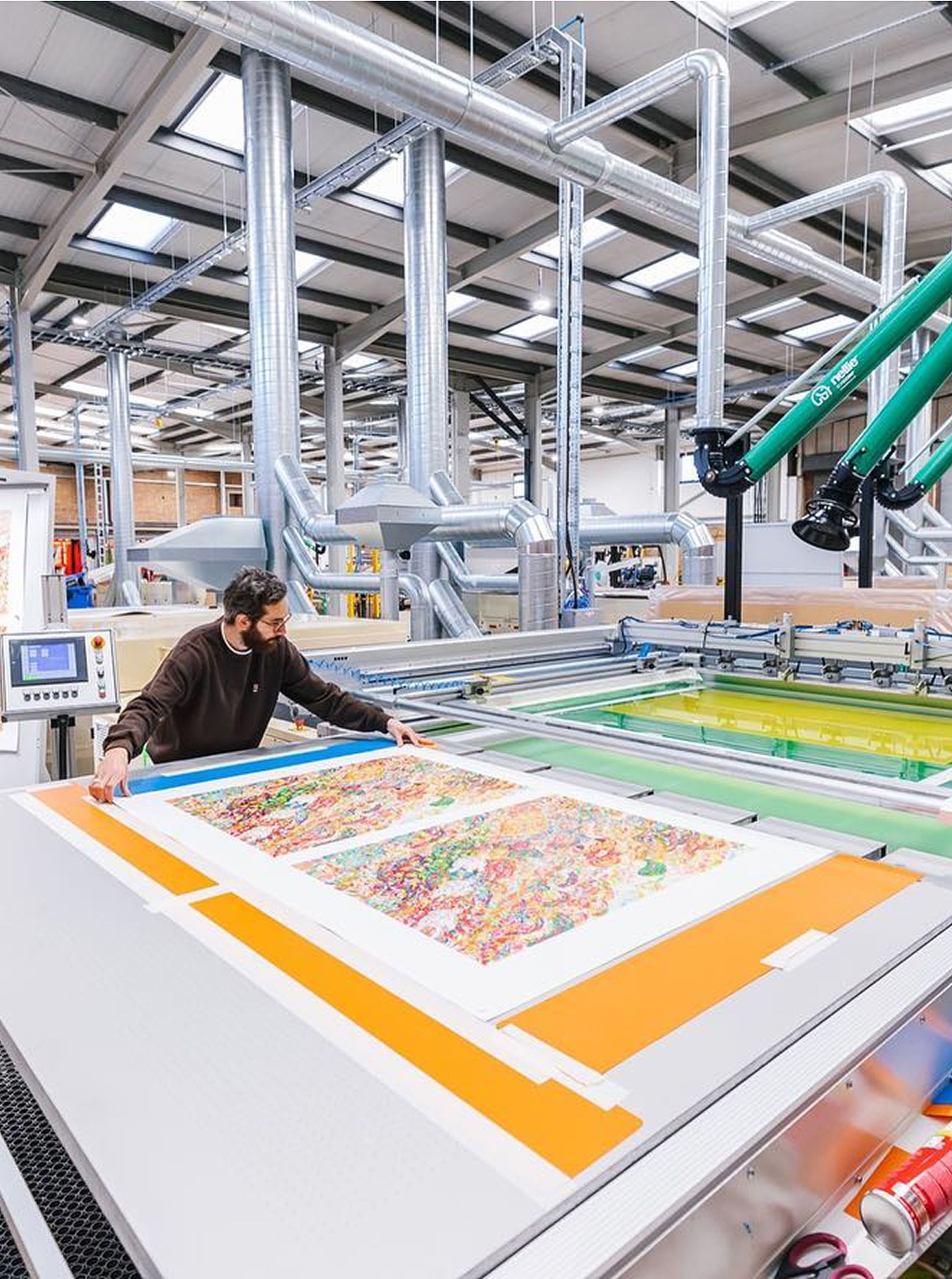 A person operates a large industrial printer, producing colorful prints in a factory setting with visible ventilation pipes and machinery.