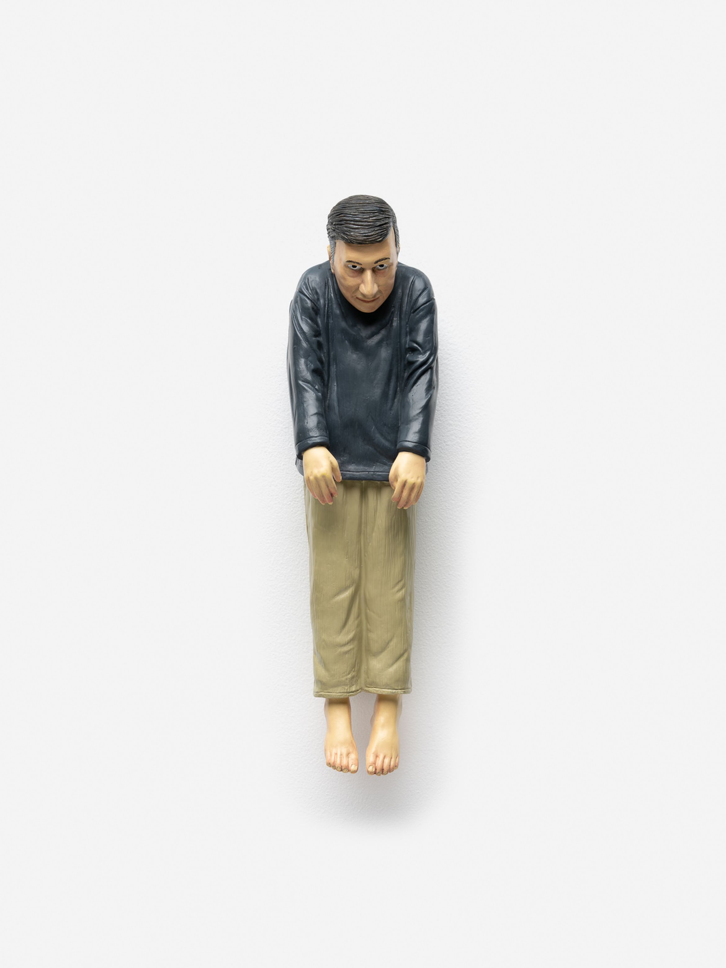 Sculpture of a man in a black shirt and beige pants, hanging with arms extended downward, against a plain white background.