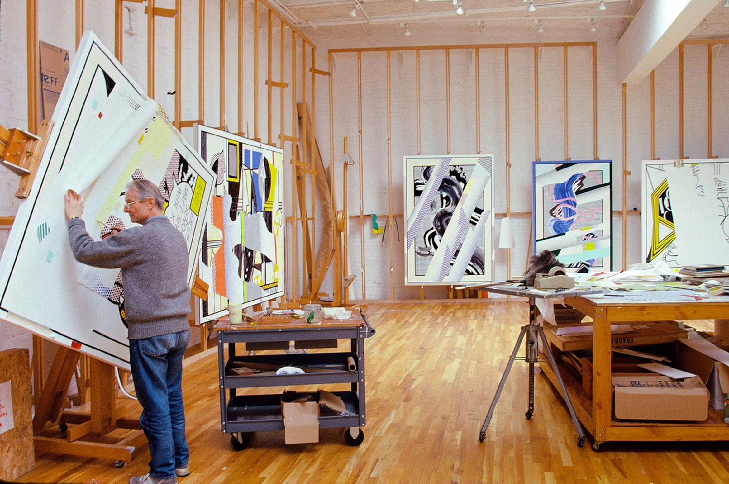 Artist Roy Lichtenstein in a spacious studio filled with colorful abstract paintings, surrounded by art supplies and a wooden floor.