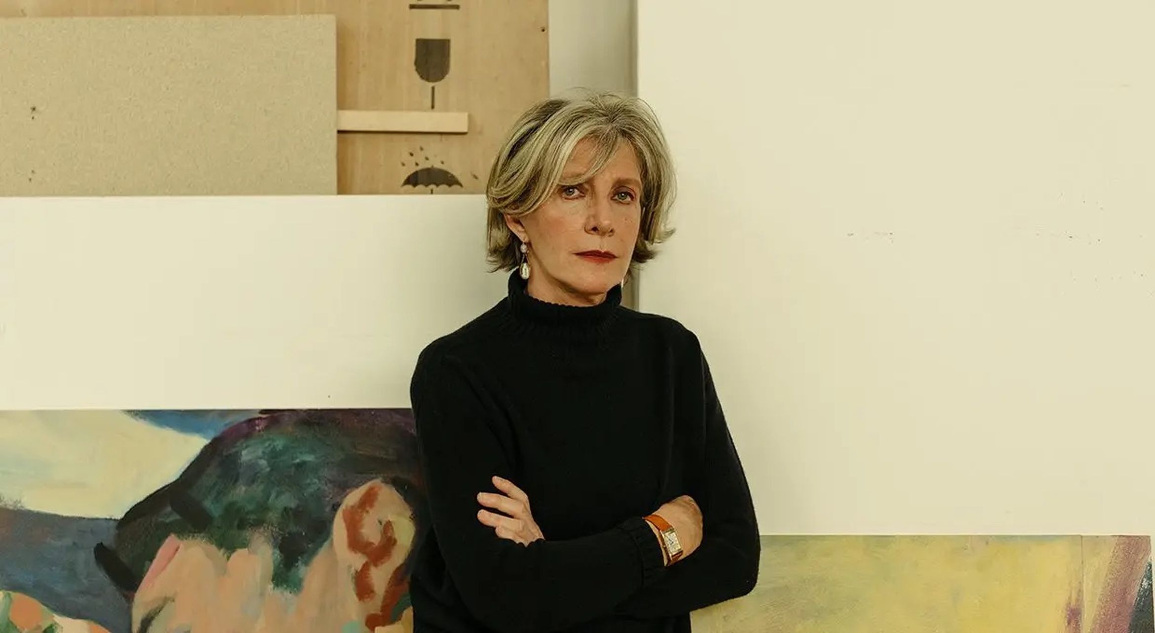 A woman with short blonde hair and a black sweater stands with folded arms in front of abstract artwork.