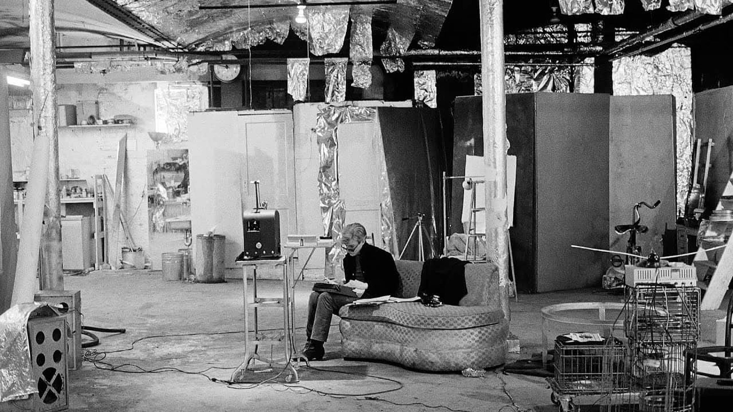 Artist Andy Warhol sitting on a couch in an industrial studio filled with various equipment, artworks, and reflective materials hanging from the ceiling.