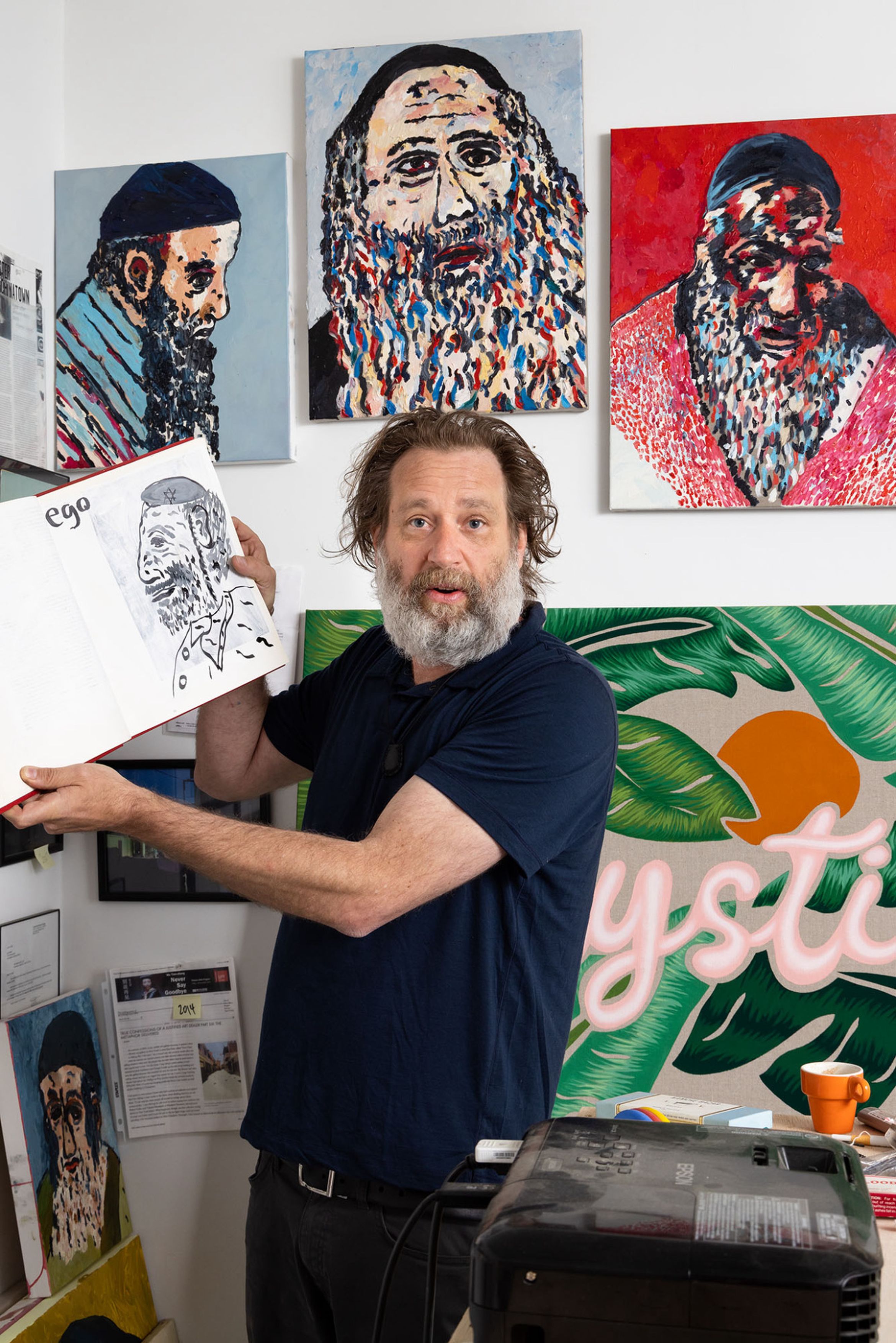 Man with a beard holding a sketchbook, surrounded by colorful portraits on a wall.