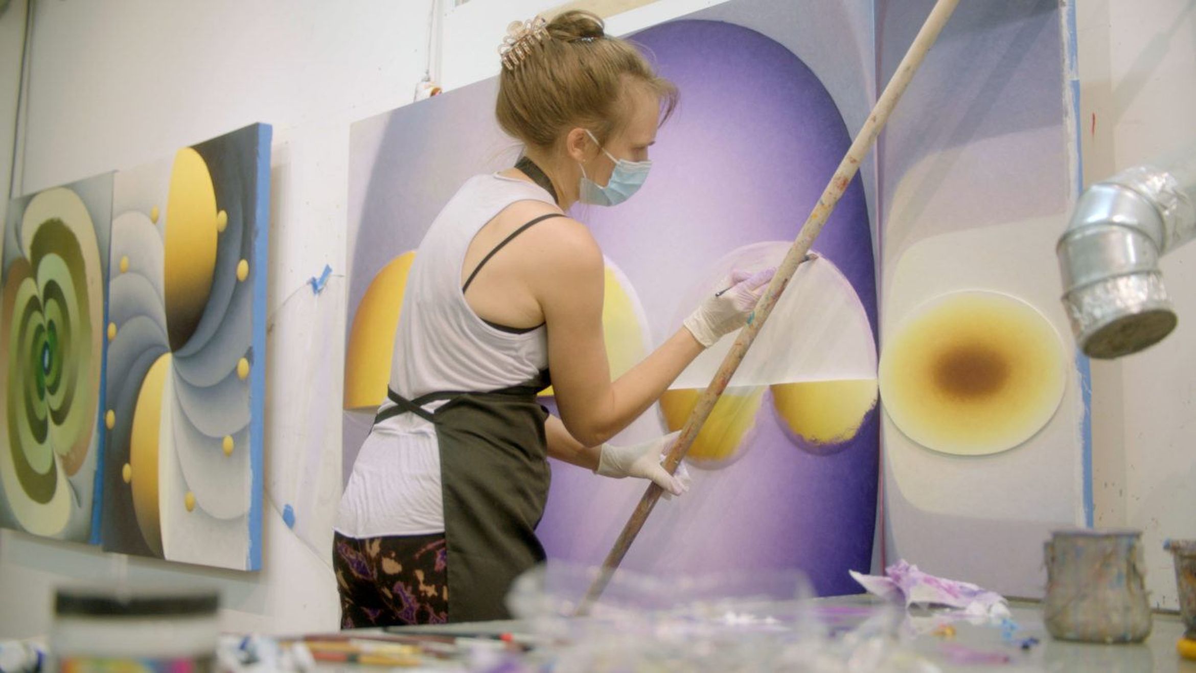 An artist wearing a mask and apron paints a colorful abstract design on a large canvas in a studio, surrounded by vibrant artworks and paint supplies.