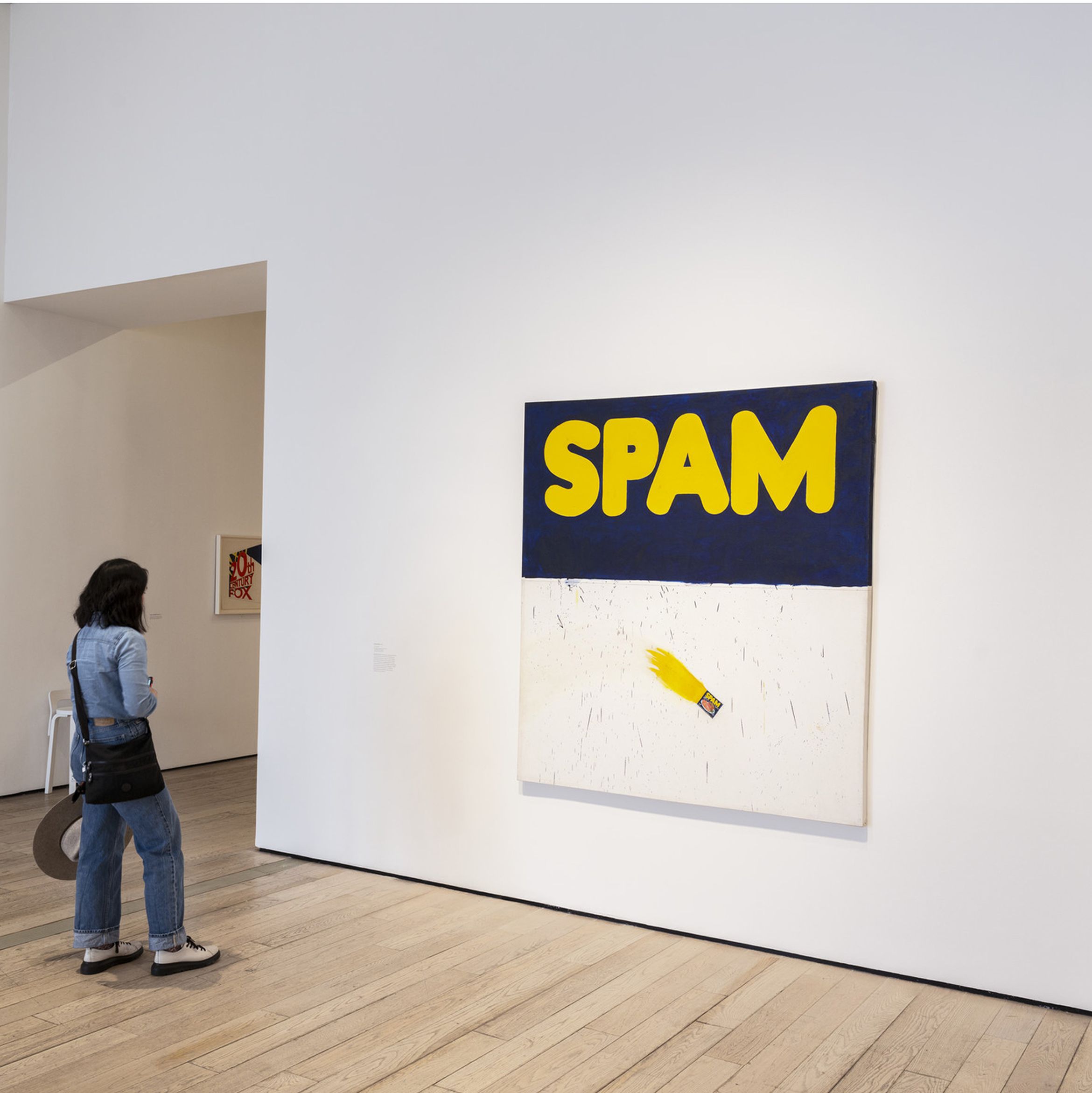 Person in denim outfit viewing a large artwork with the word "SPAM" in bold yellow letters on a blue and white background in a gallery.